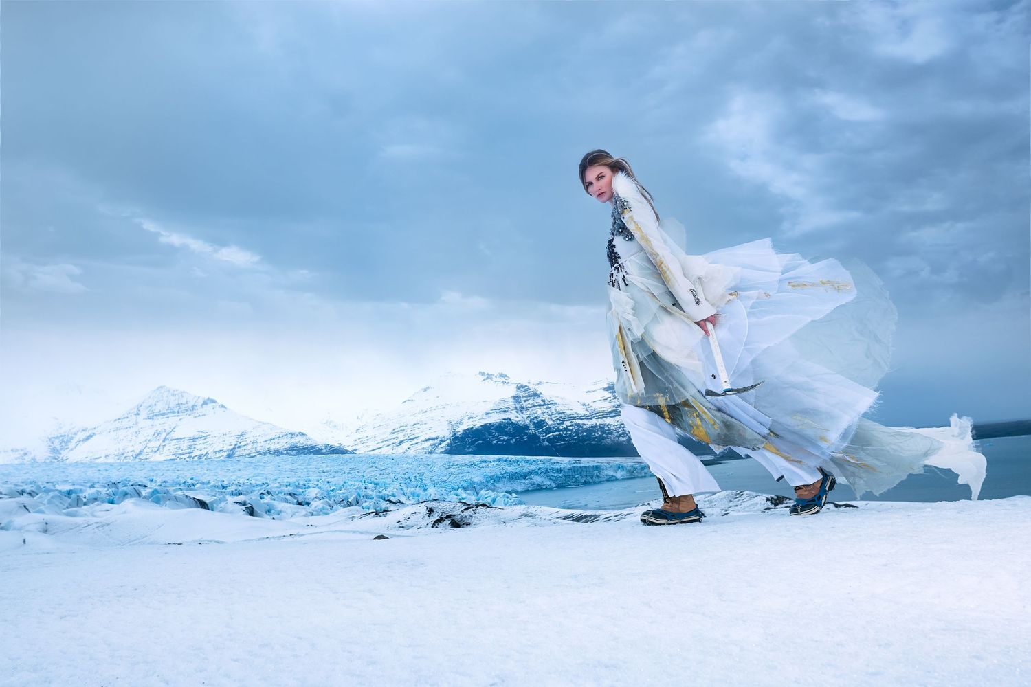 A person in flowing white dress poses dramatically against a snowy mountain landscape with cloudy blue skies.
