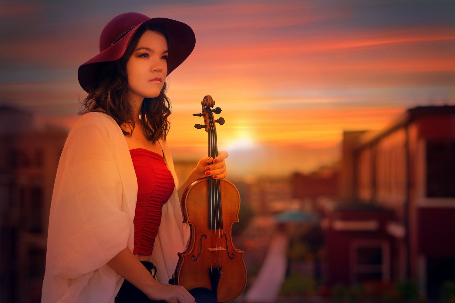 A violinist in a red dress and white shawl poses with their instrument against a vibrant sunset sky.