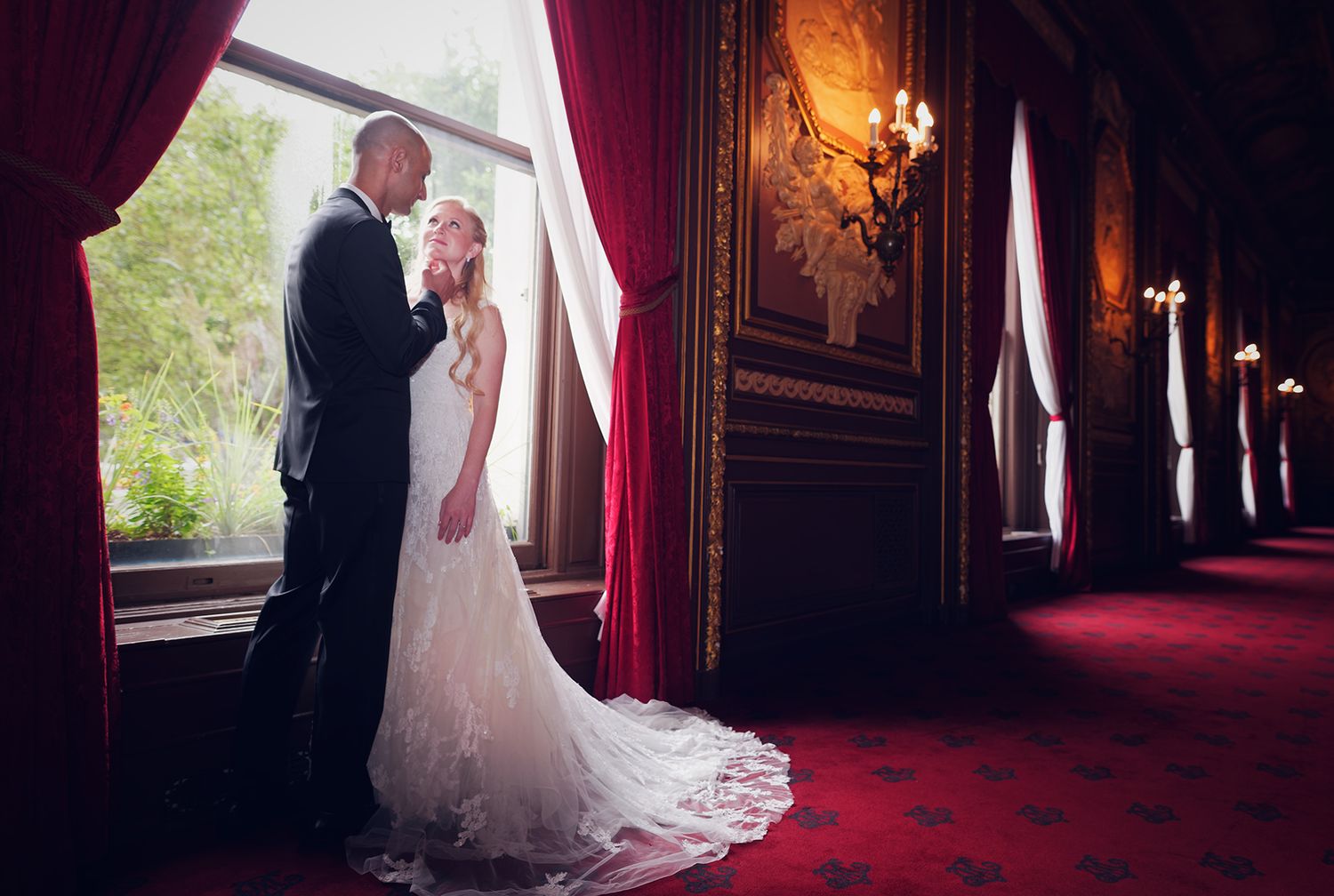 Elegant wedding couple stands by window in ornate historic building with red carpet and candle sconces.