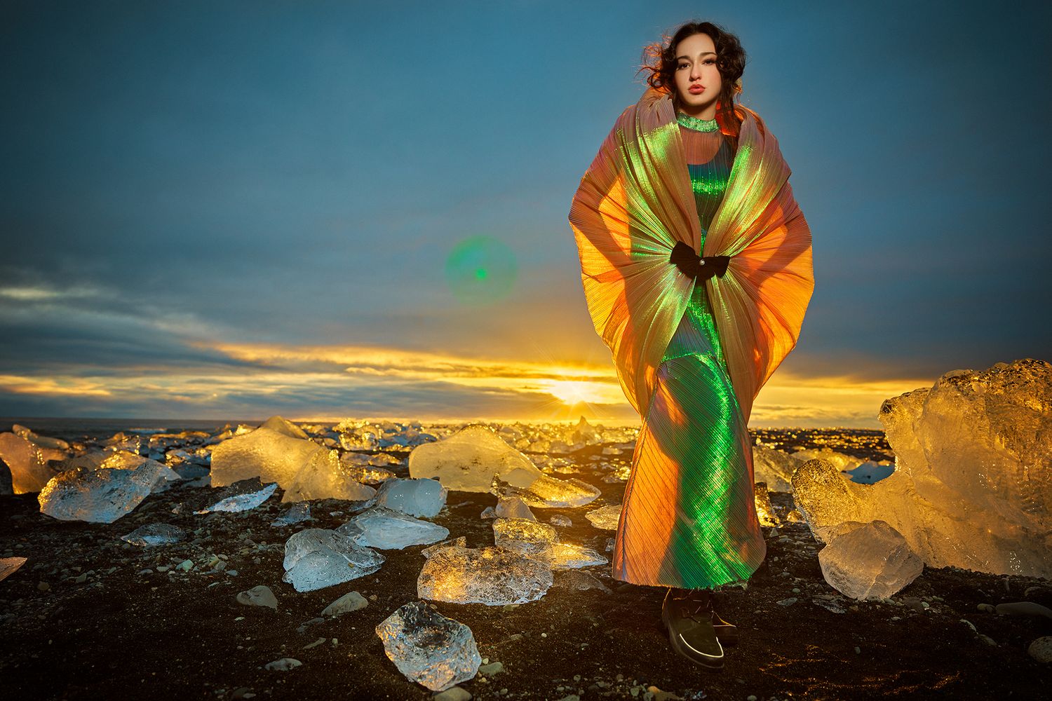 A figure stands among ice formations at sunset wearing vibrant green and orange flowing fabric against a dramatic sky.