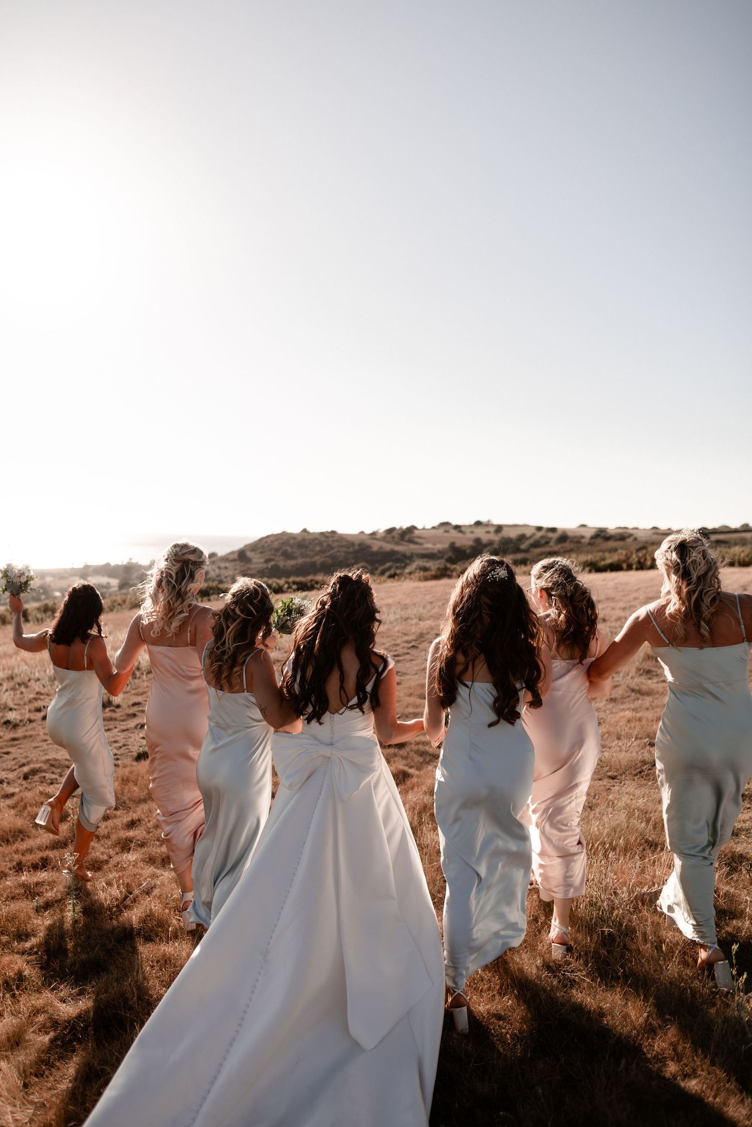 A group of people in white dresses pose together on a hillside during golden hour in a panoramic shot.