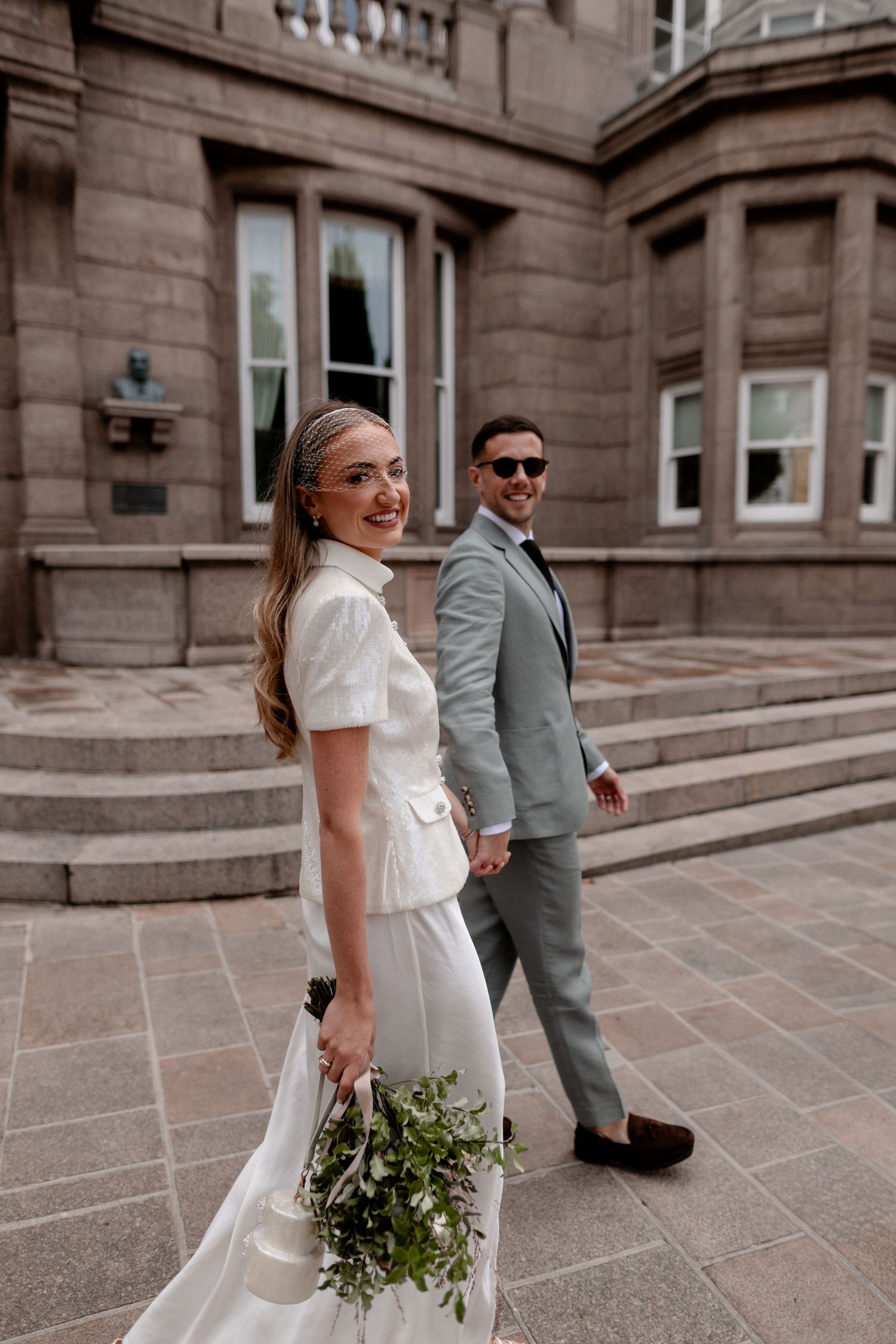 Bride in white dress holding bouquet walks with groom in gray suit outside a stone building.