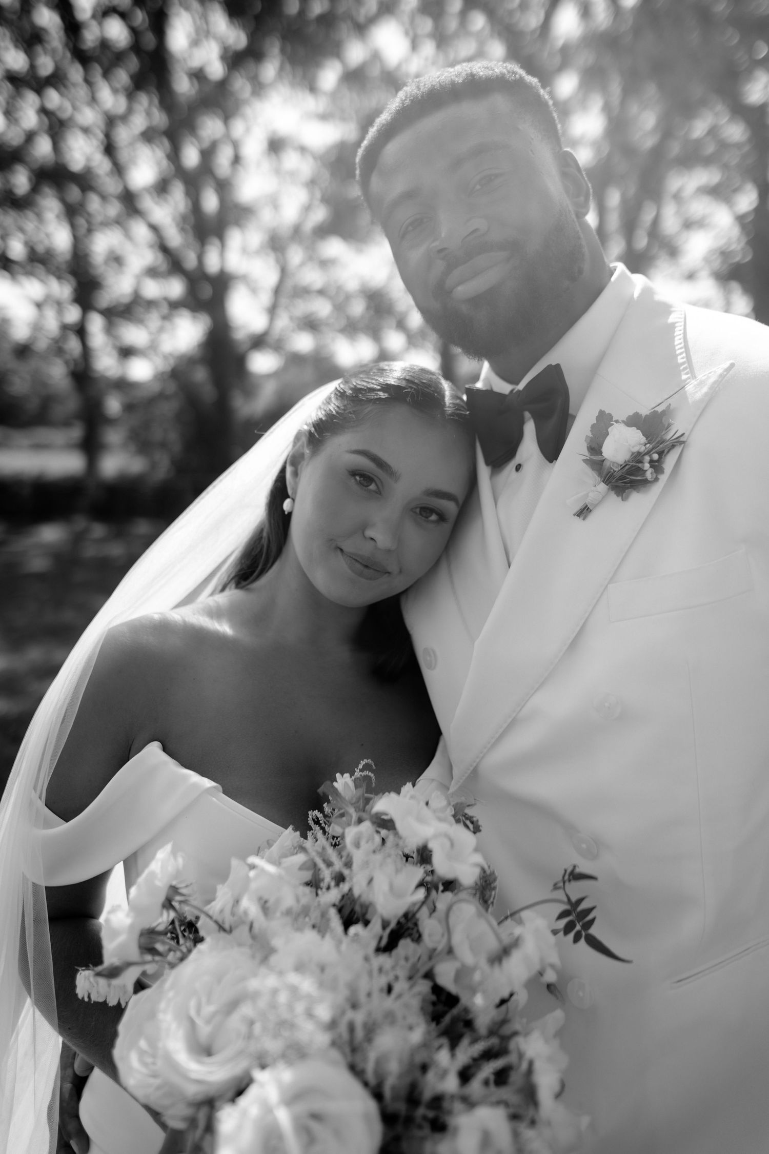 Black and white wedding portrait of bride holding bouquet leaning on groom in white tuxedo outdoors.
