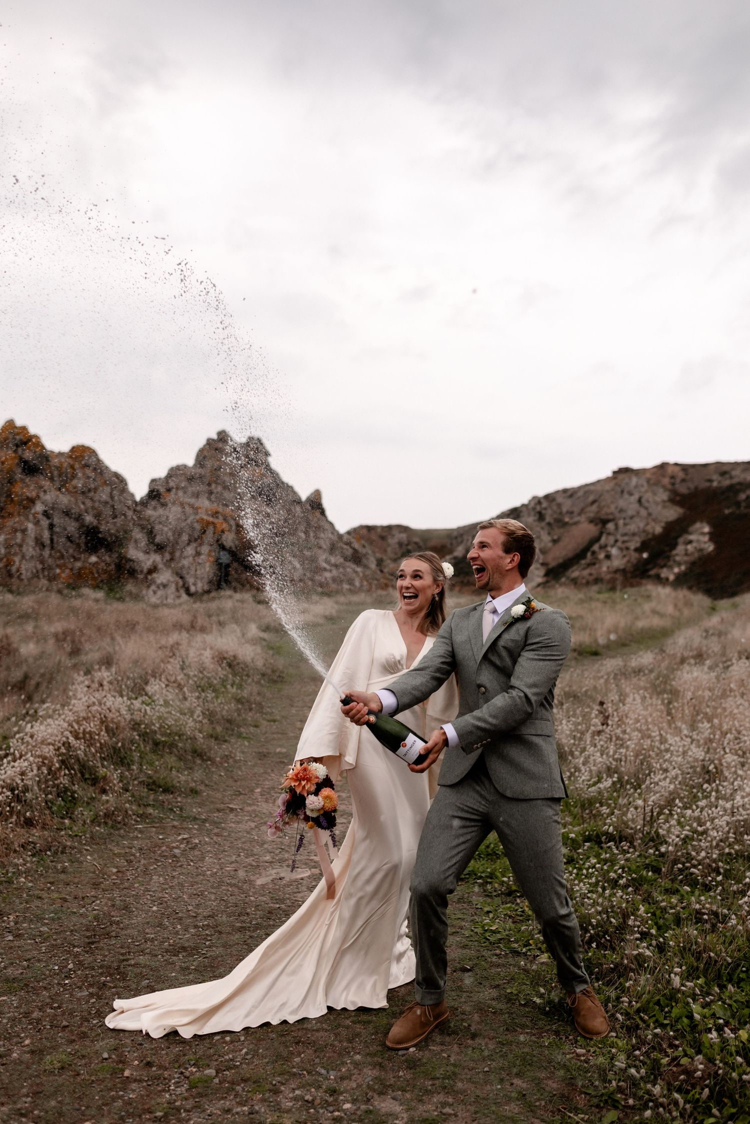 Joyful bride and groom popping champagne outdoors on a rocky trail, celebrating their wedding day.