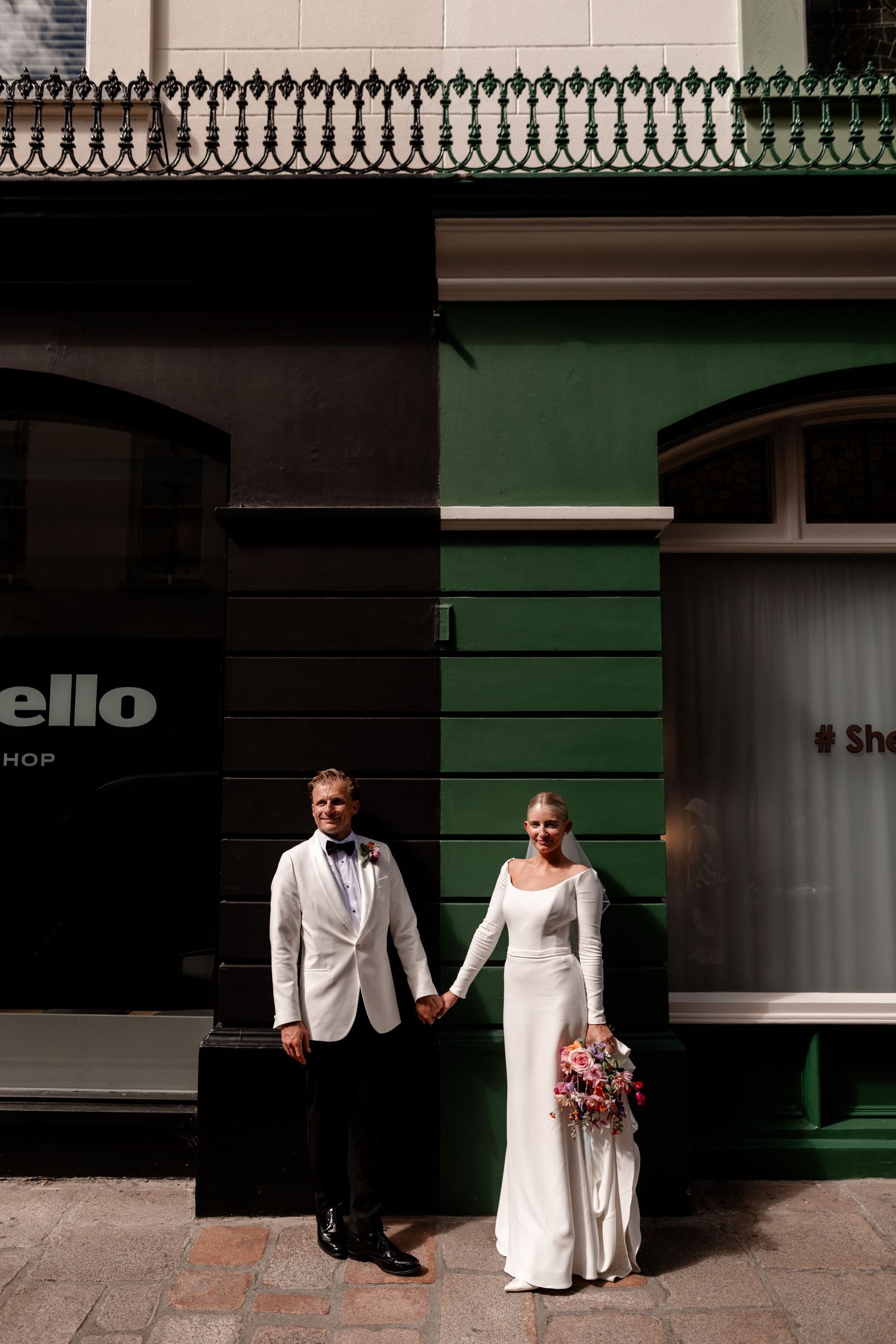 Bride and groom holding hands in front of a green building, bride holding a colorful bouquet in a sleek white gown.