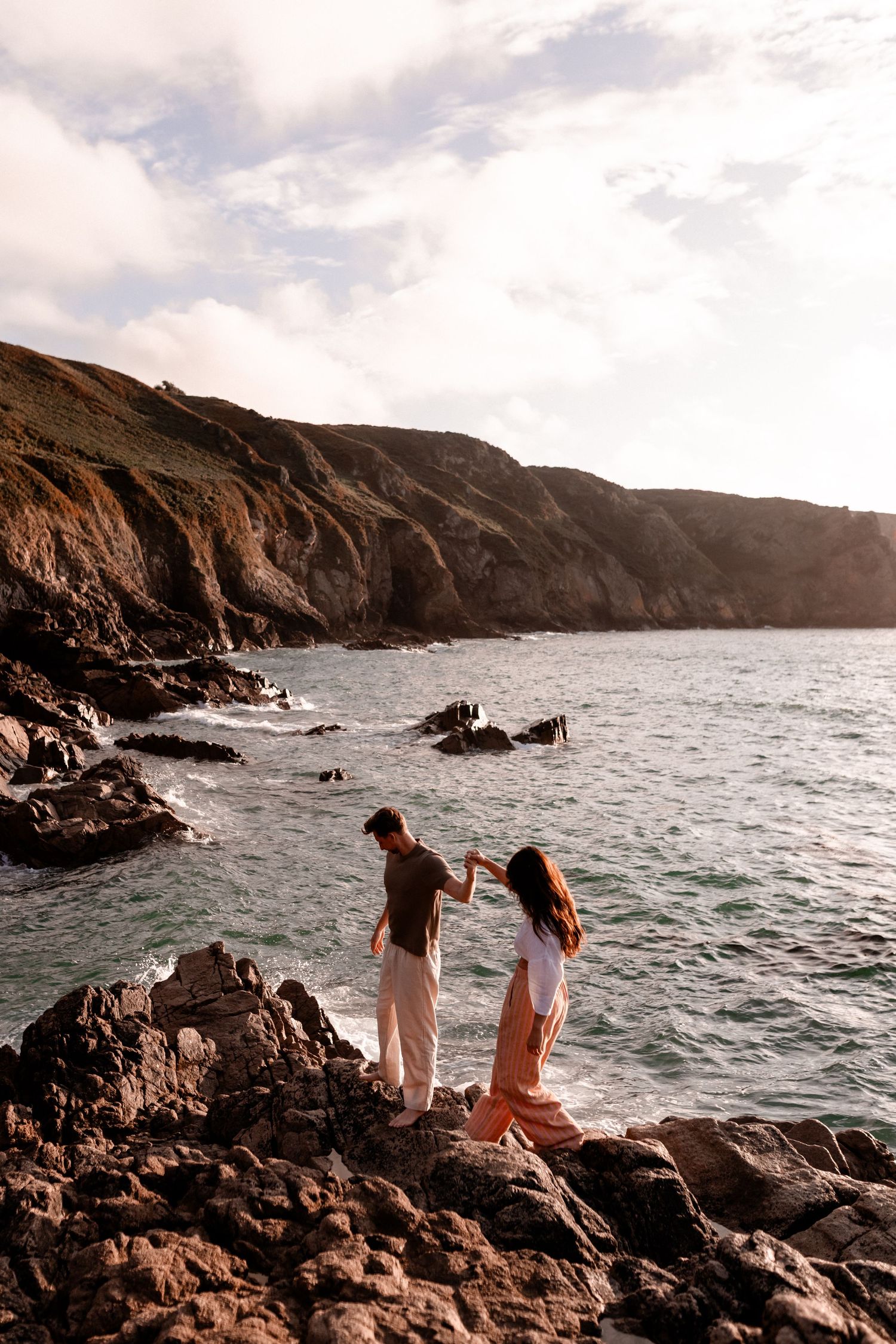 Couple holding hands on rocky coastal cliffs with turquoise ocean waves and dramatic hillside backdrop.