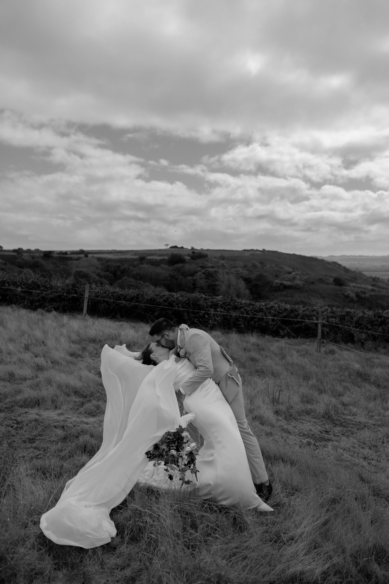 Black and white photo of a couple sharing a romantic dip kiss in a grassy field, bride's veil flowing in the wind.