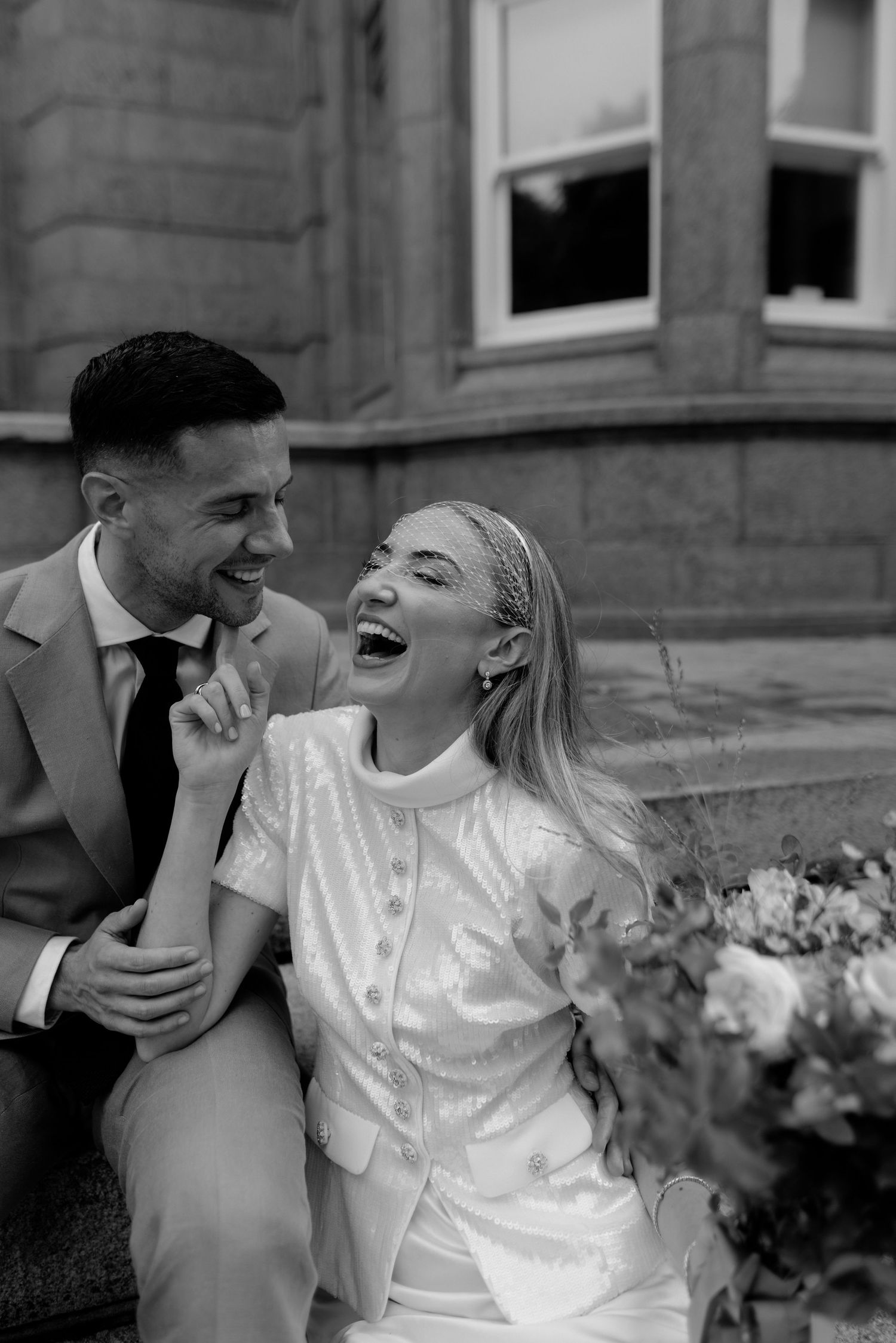 Black and white photo of a laughing bride in white dress and groom in suit sitting together on steps outside a building.