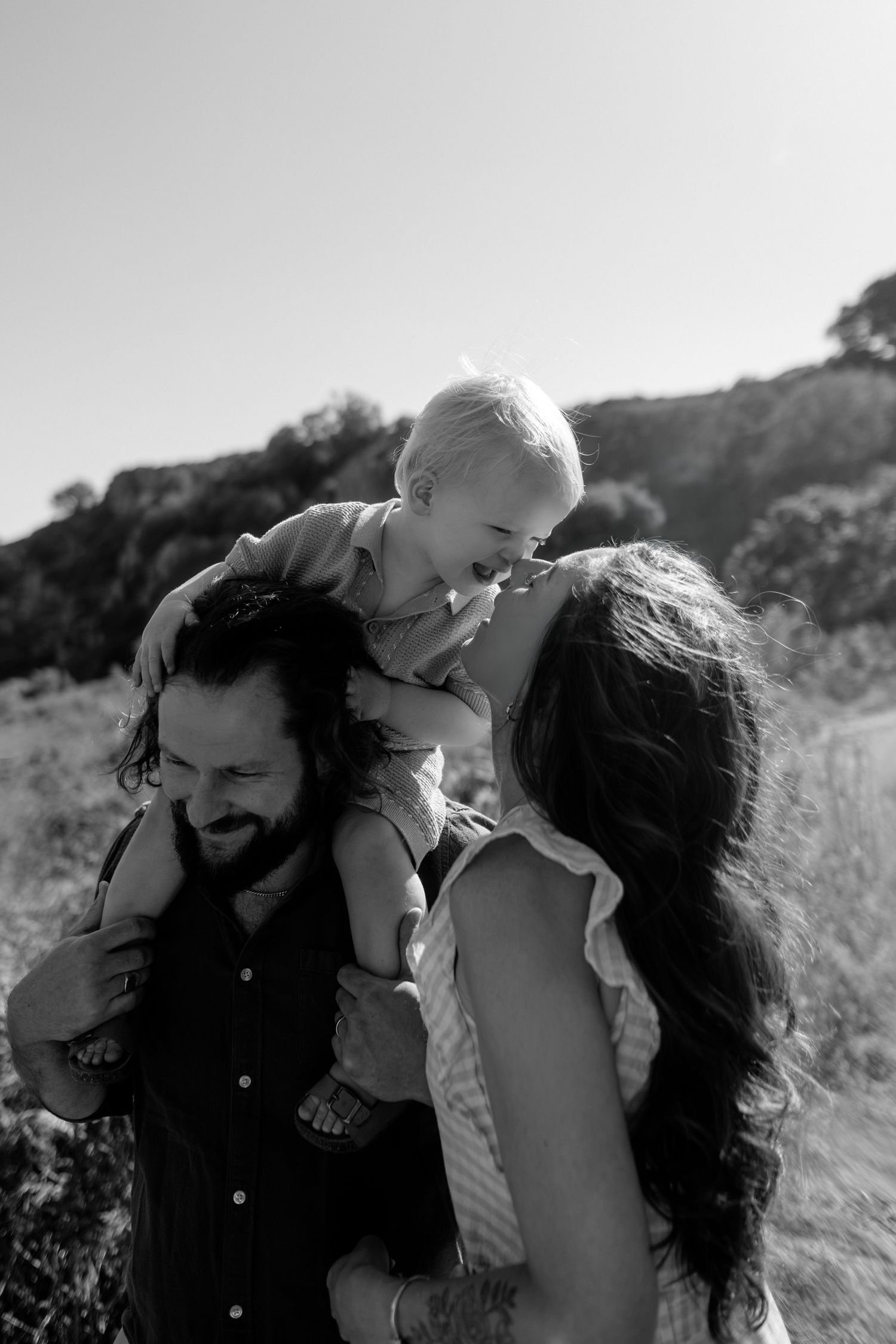 Black and white photo of a bearded father with toddler on shoulders, smiling with long-haired mother outdoors.