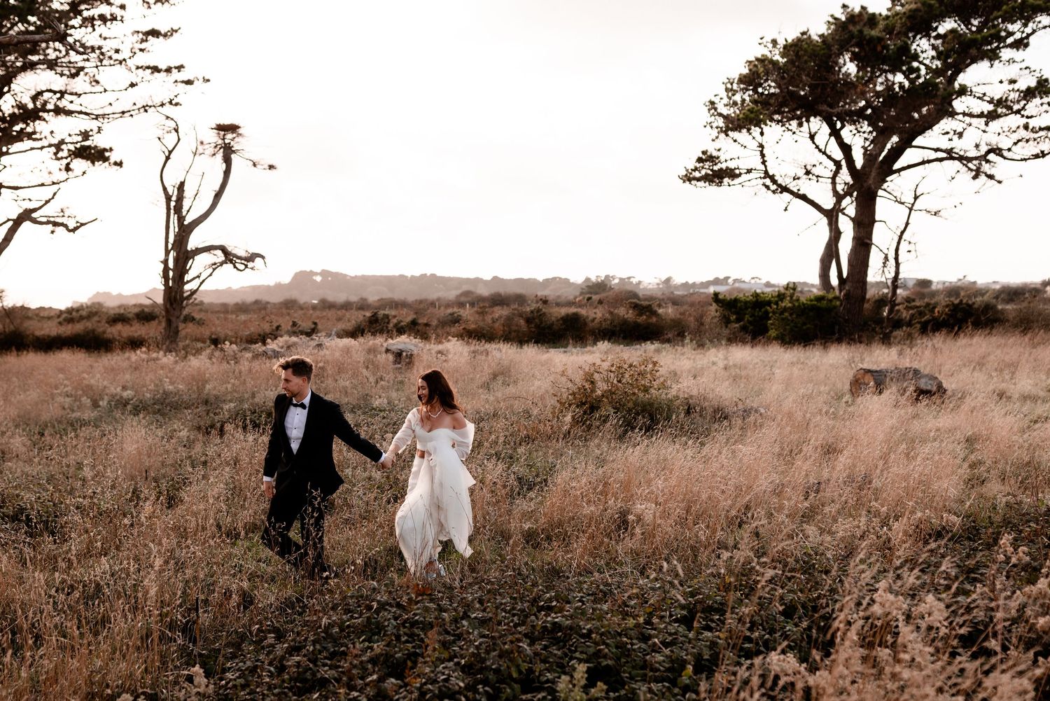 Newlyweds run joyfully through a rustic field with windswept trees in the background.