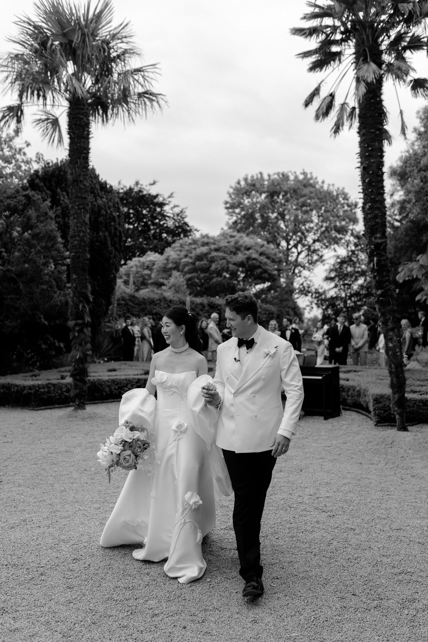 A wedding couple walks along a palm-lined garden path in elegant attire during their special day.