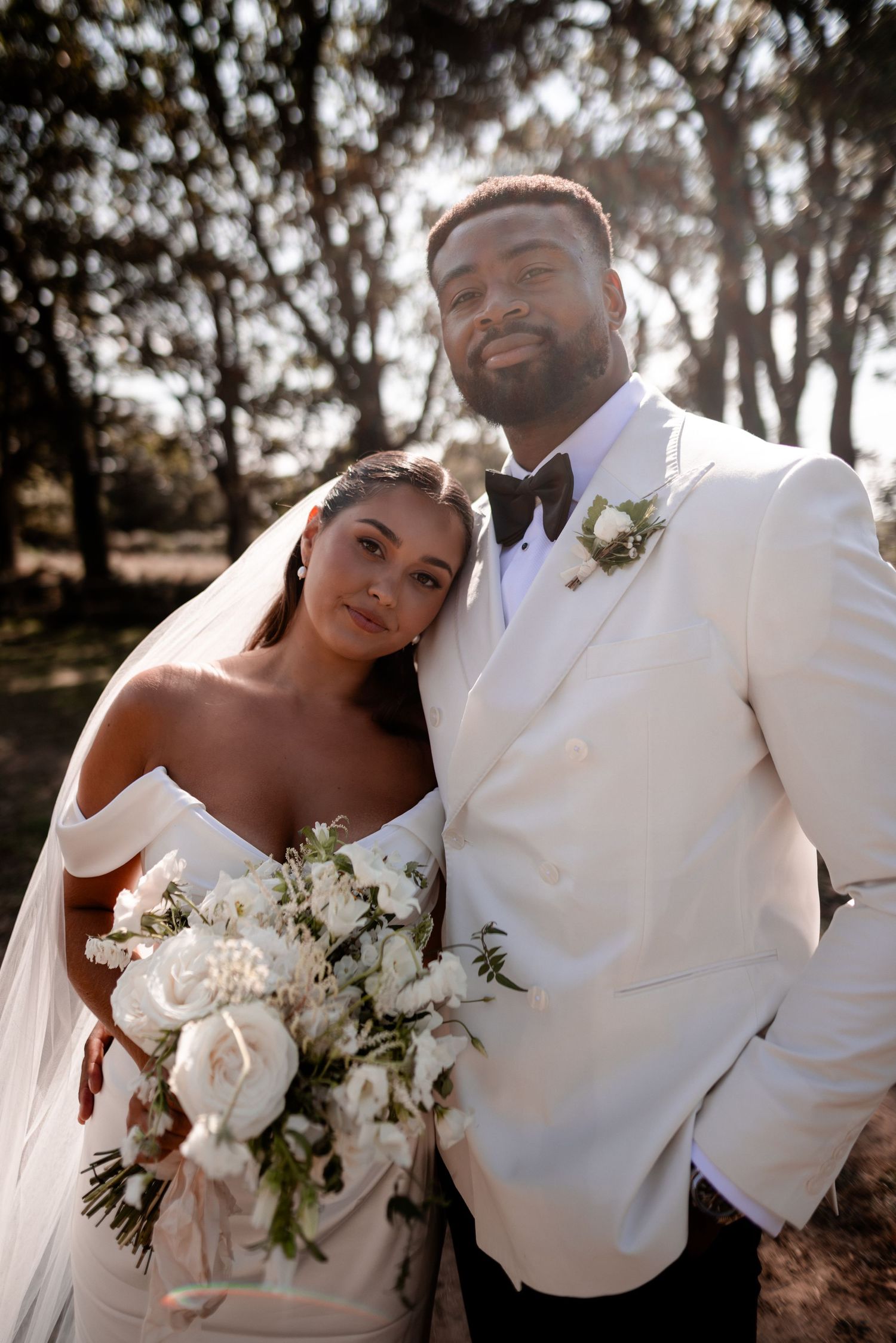 Bride and groom pose outdoors in wedding attire, she holds white floral bouquet, he wears white tuxedo jacket with boutonniere.