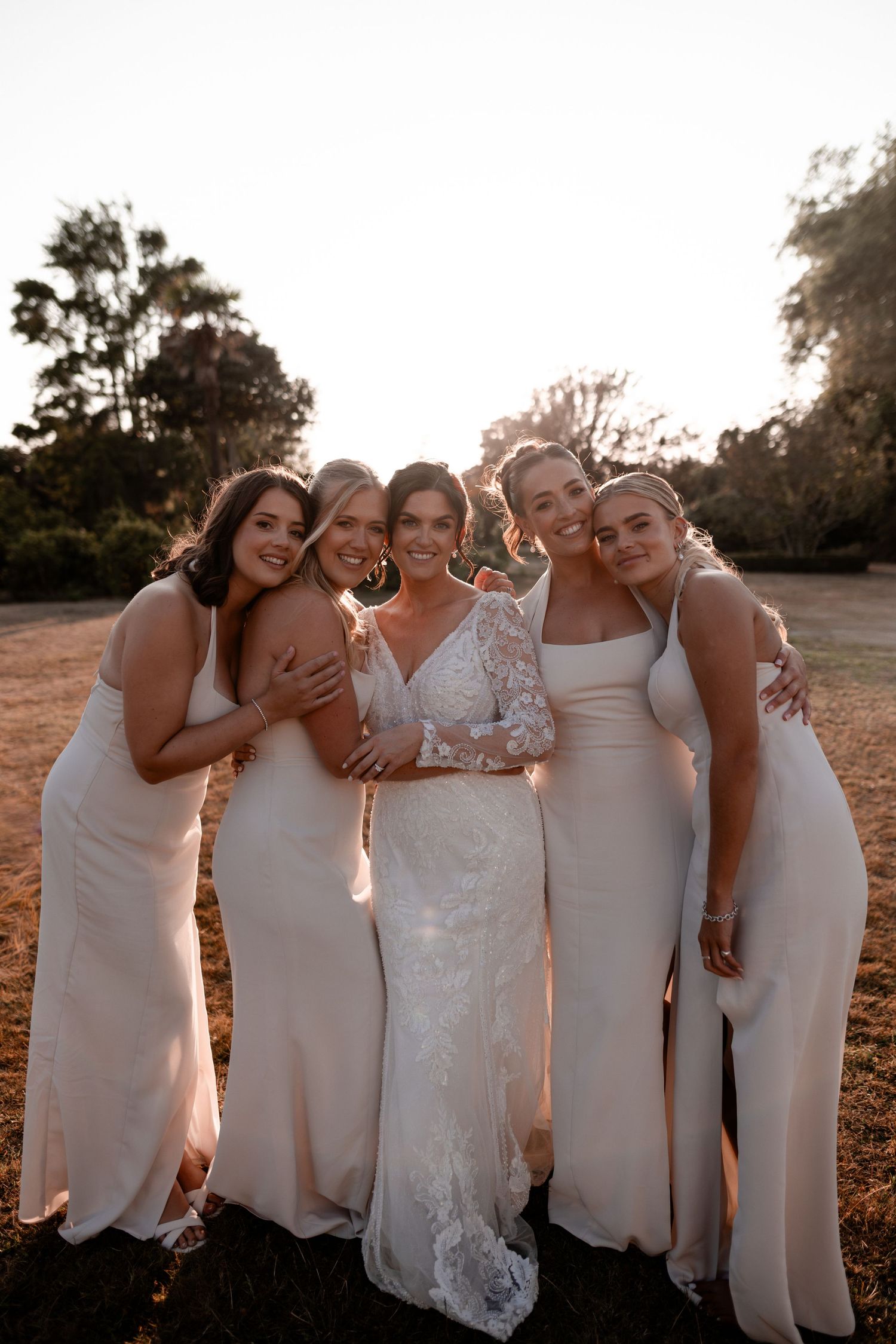 Bride in lace gown poses with four bridesmaids in ivory dresses at golden hour outdoor wedding ceremony.