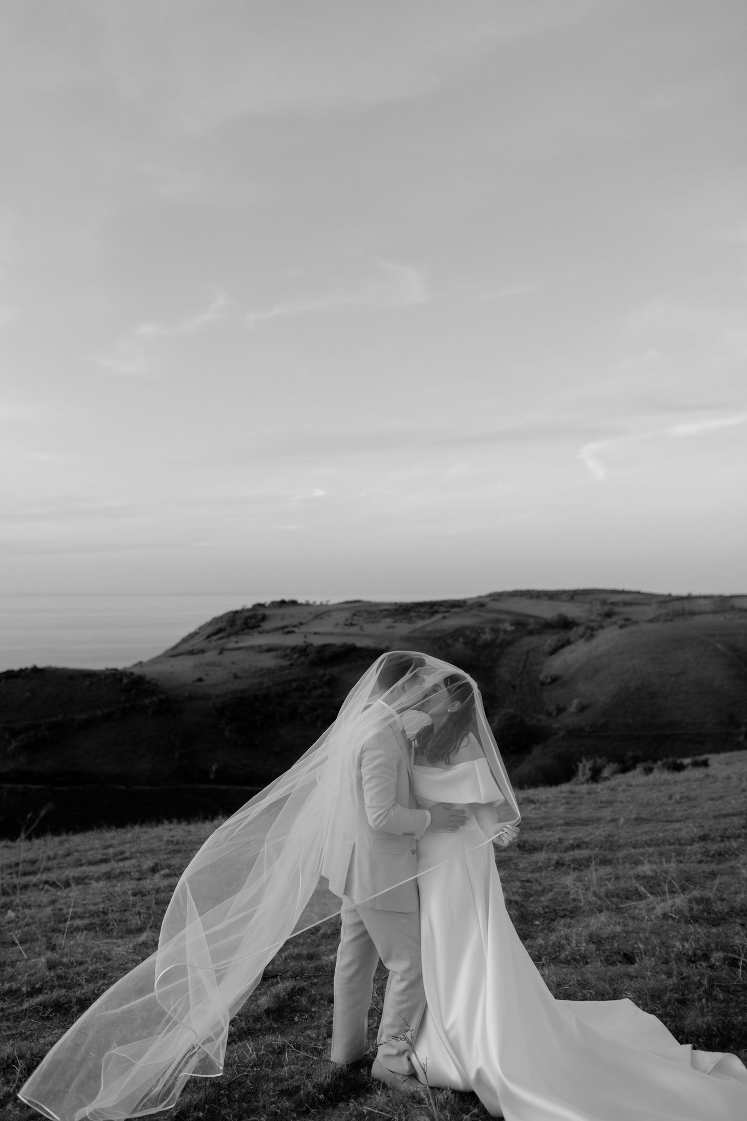 Black and white photo of bride and groom kissing under flowing veil on scenic hilltop at sunset.