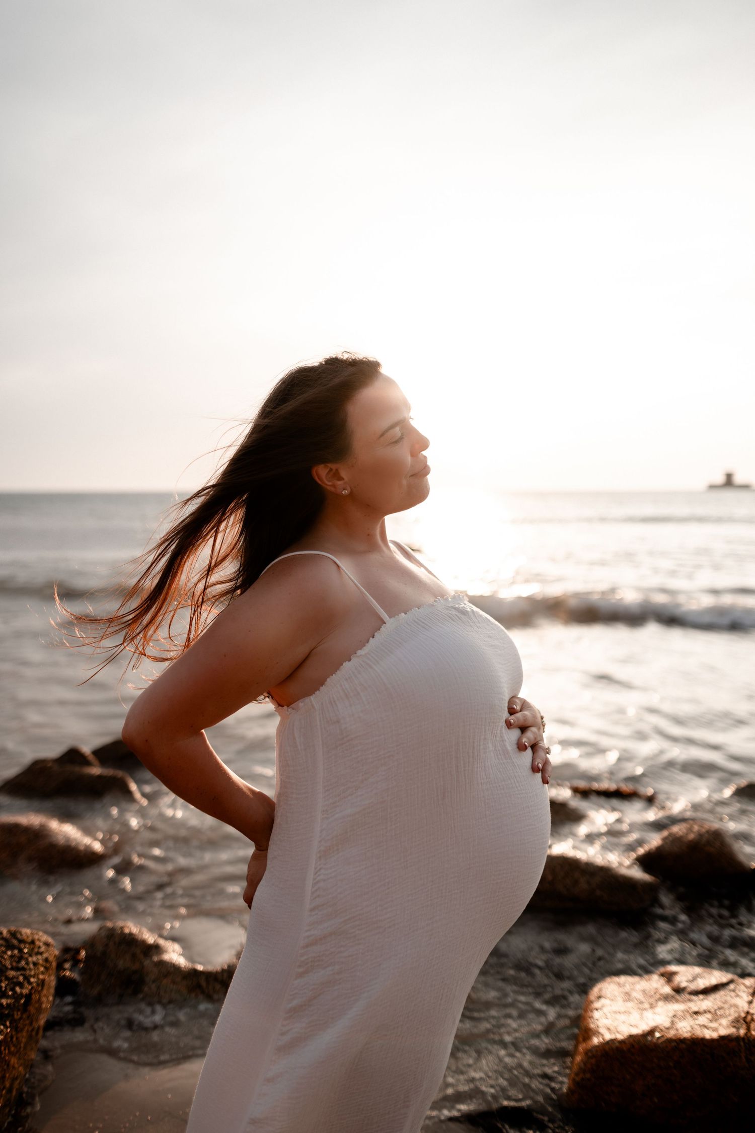 Pregnant woman in white dress holding belly on rocky beach at sunset with ocean waves behind her.