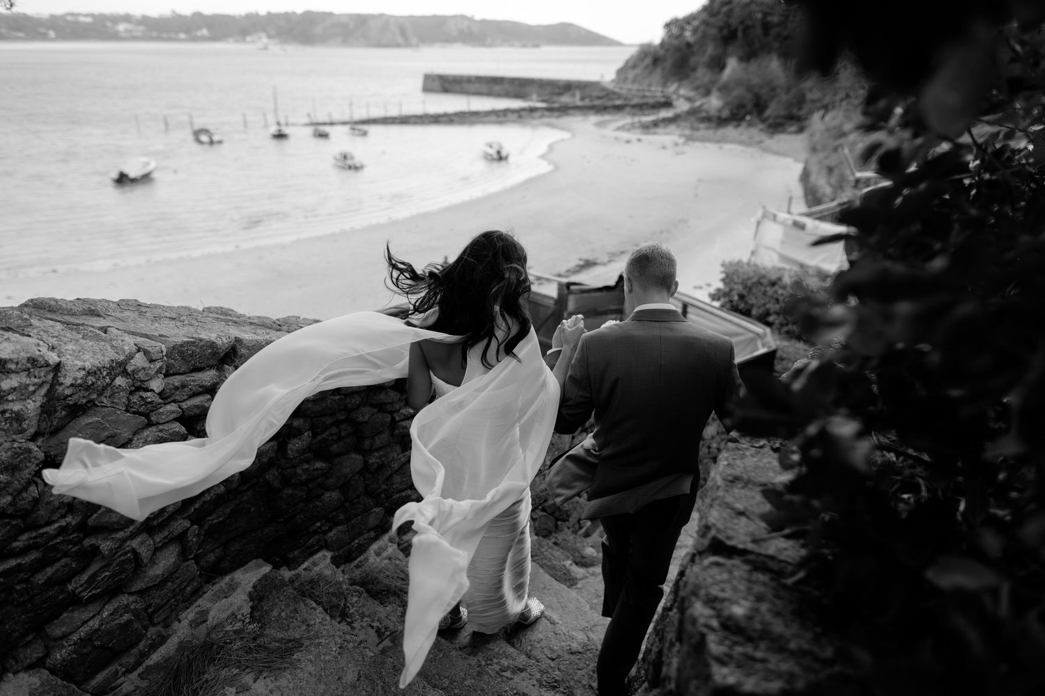 Black and white photo of a bride and groom walking up stone steps toward a scenic coastal harbor with boats.