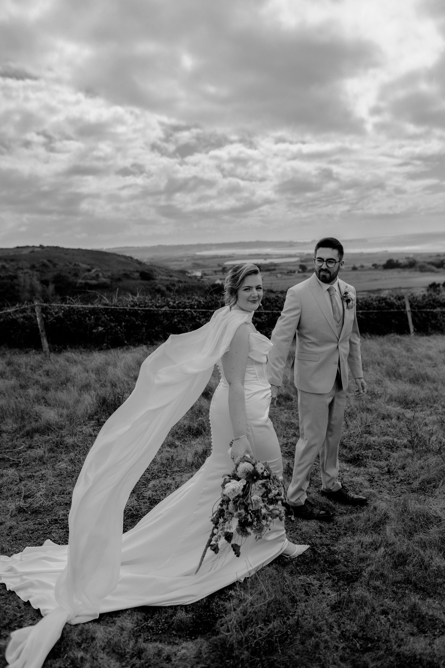 Black and white photo of a bride and groom holding hands in a field, her veil flowing in the wind.