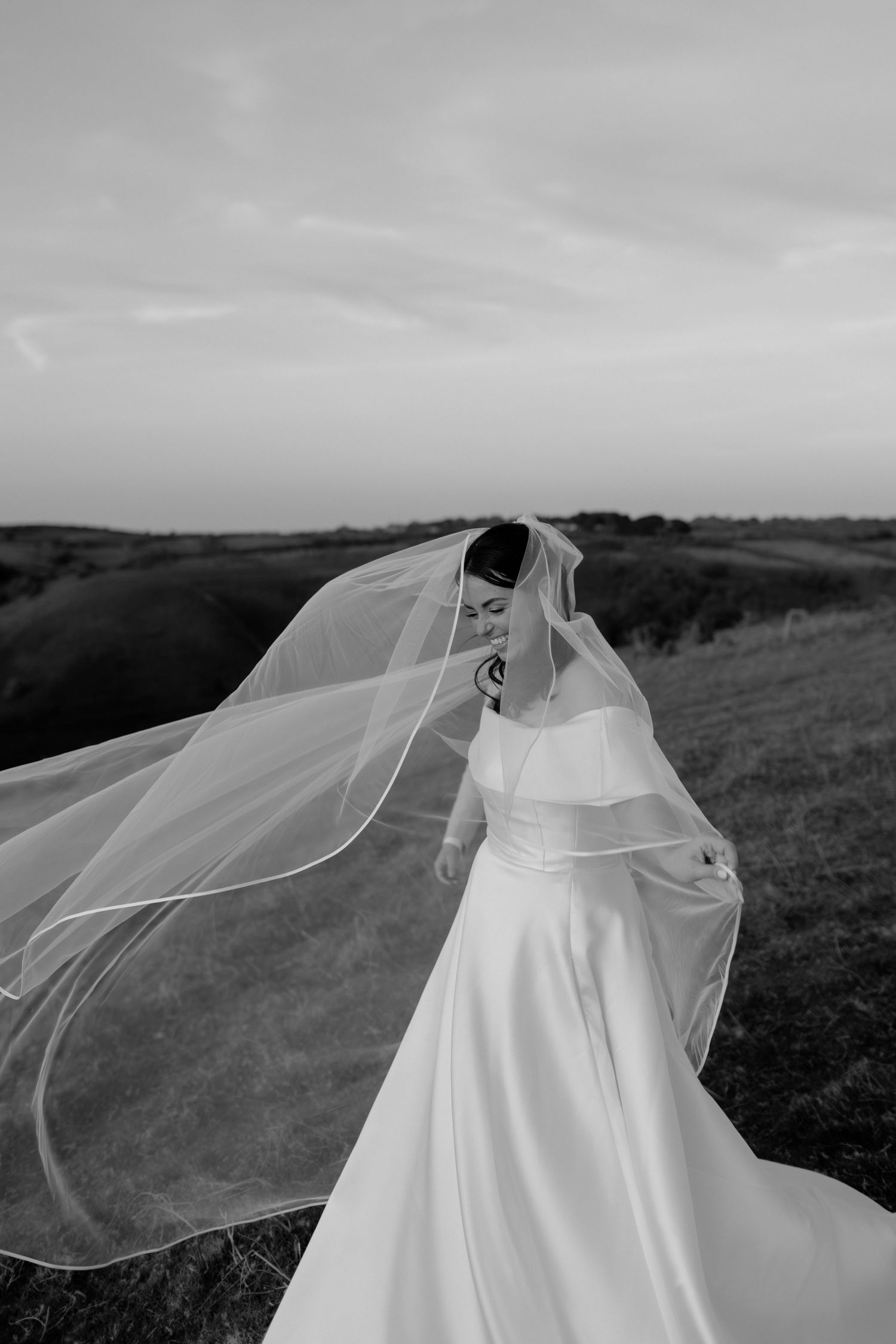 Black and white photo of a bride in a flowing white gown and veil blowing in the wind on a scenic hillside.