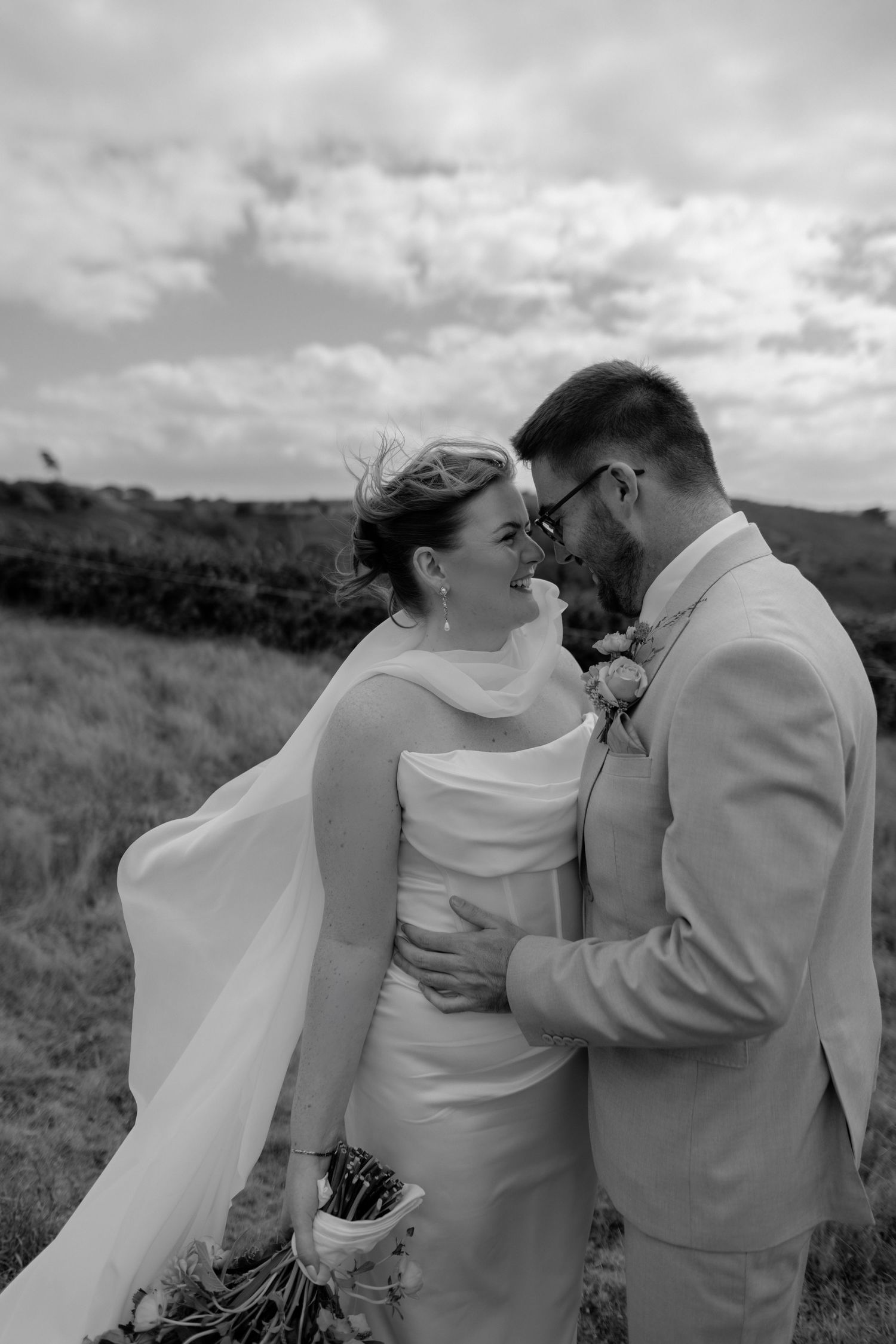 Black and white photo of a bride and groom embracing outdoors, smiling at each other on their wedding day.