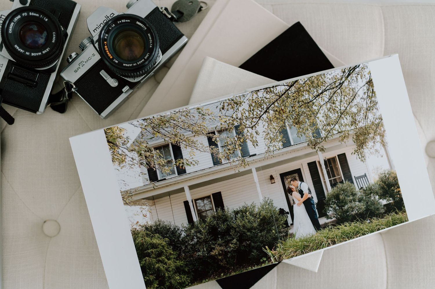 A vintage photograph of a white colonial-style house with film cameras displayed on a white surface beside it.