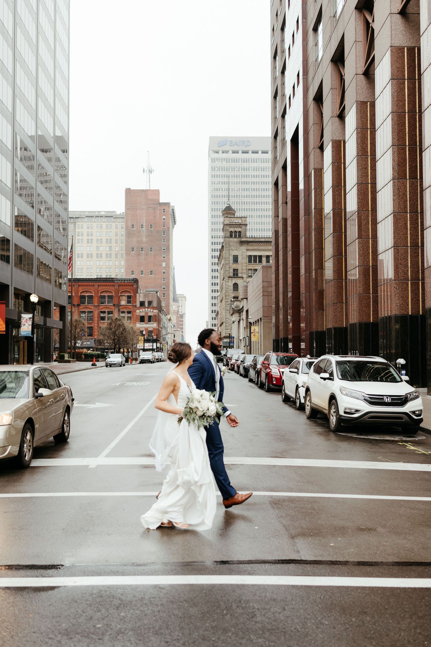 A bride in white dress and groom in navy suit crossing an urban street between tall buildings.