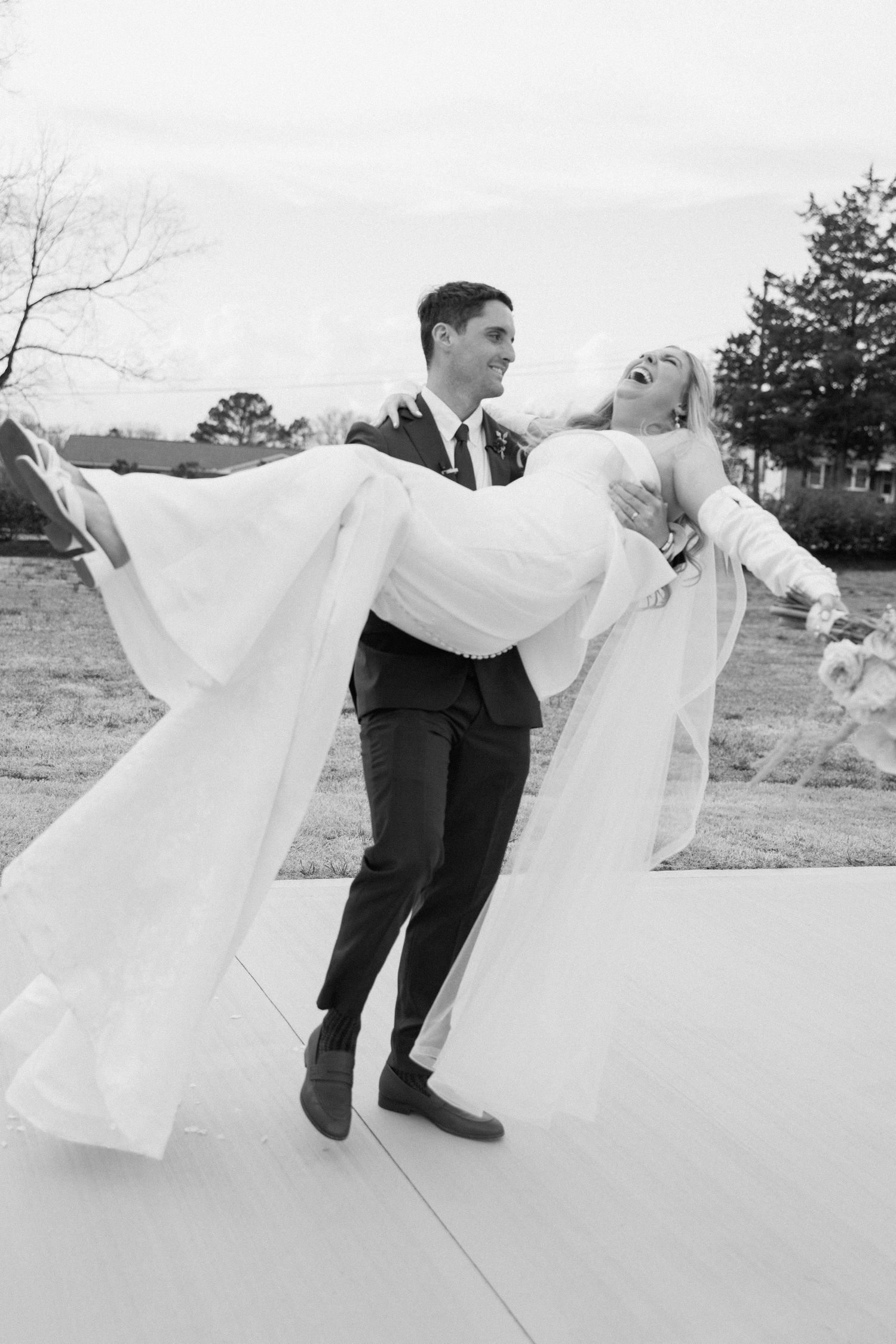 Groom carries bride in wedding dress outdoors in joyful black and white wedding photo.