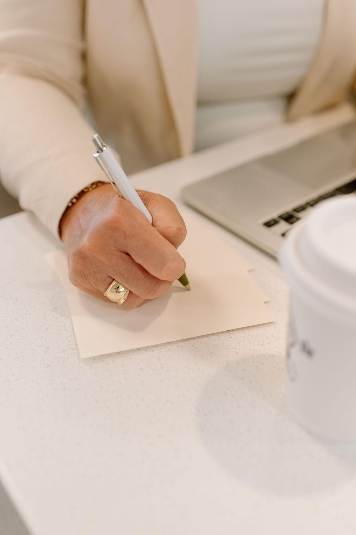 Hand with cream sweater sleeve and gold ring holding pen to write on white paper next to laptop and coffee cup.