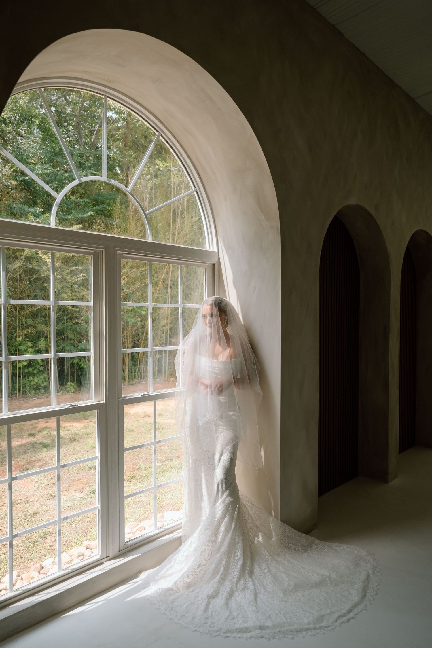 Bride in lace wedding gown and long veil stands by arched window in elegant venue.