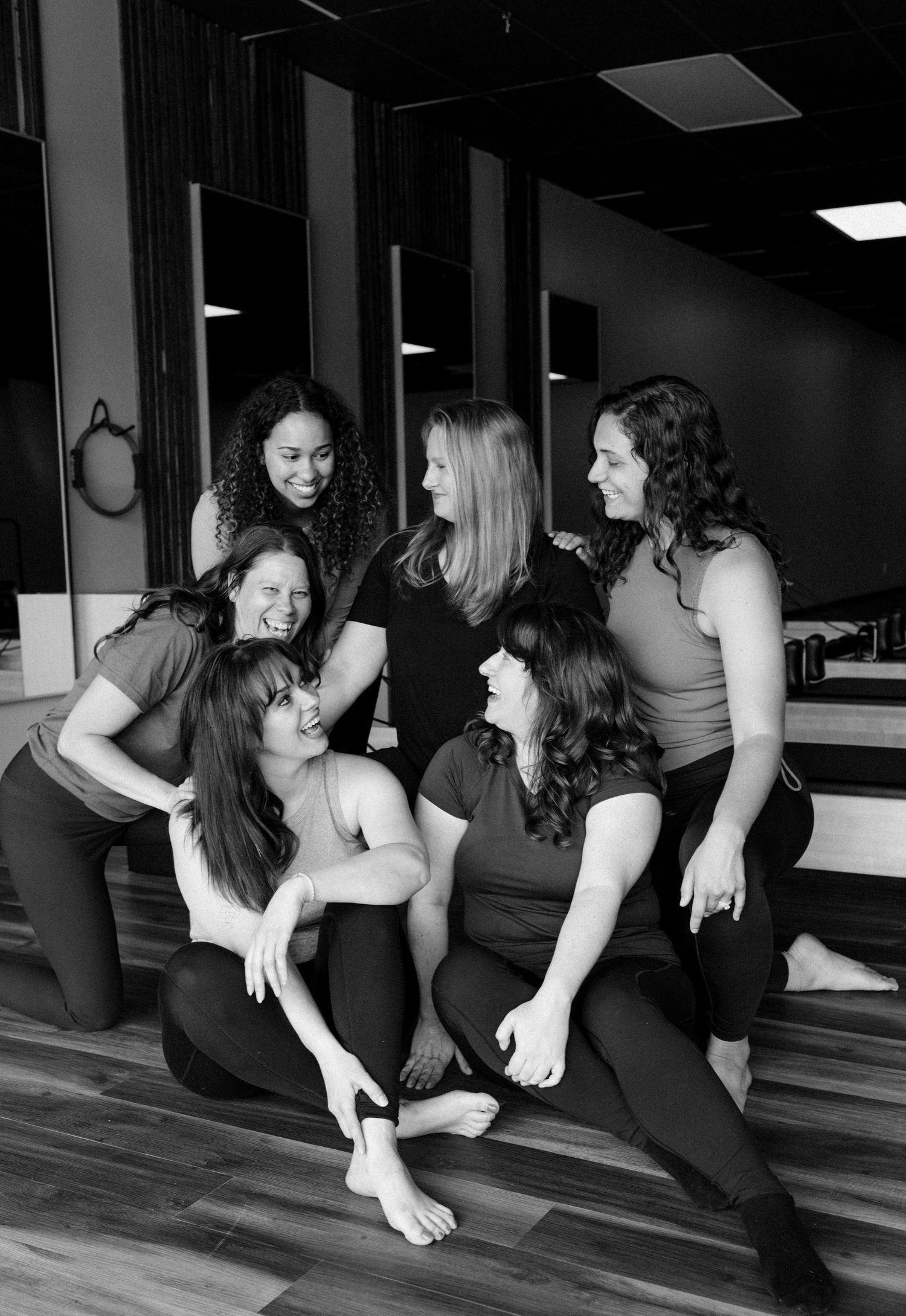 A group of six women in black activewear sit and laugh together in a dance or yoga studio.
