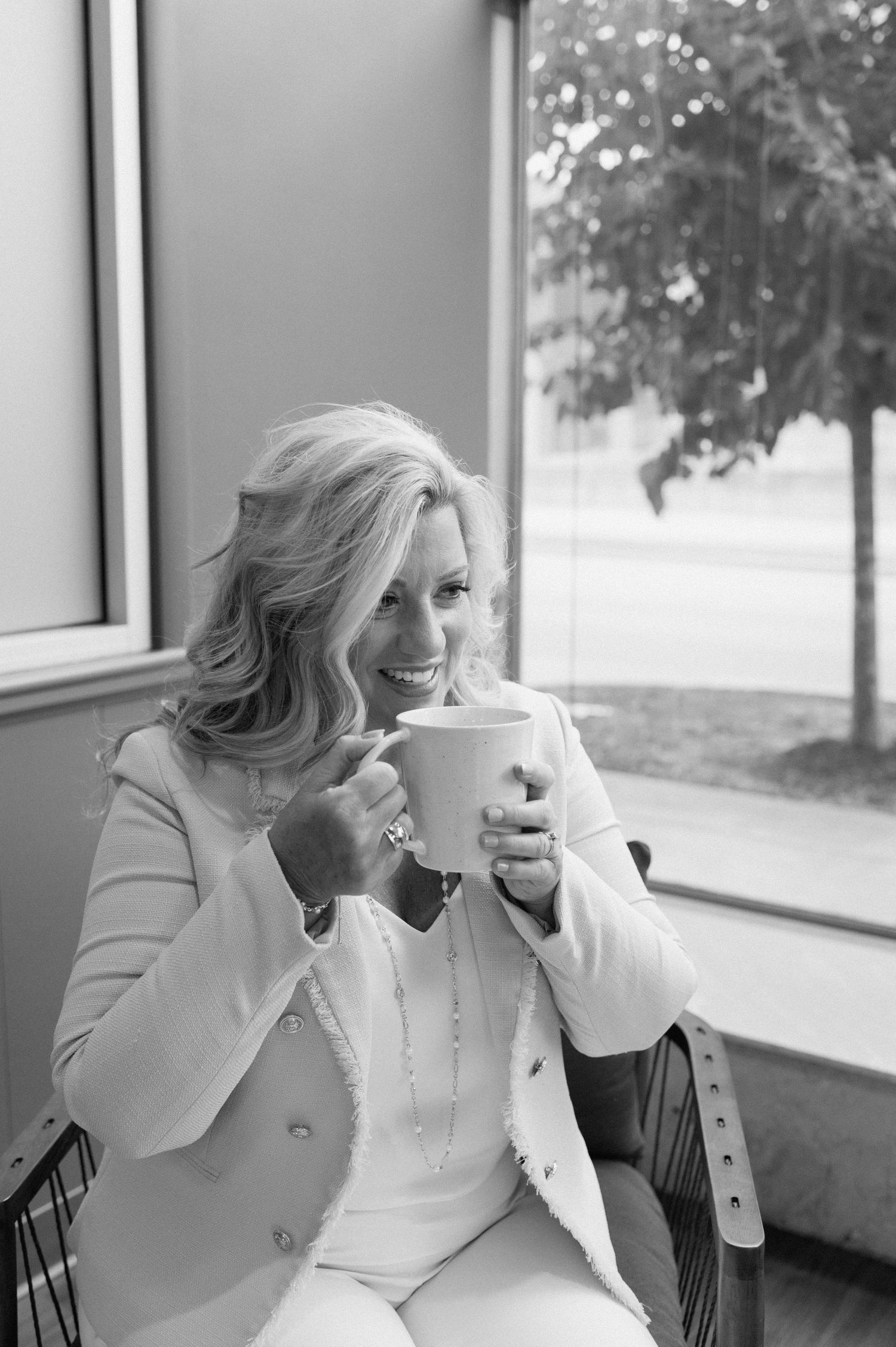 Black and white photo of someone enjoying coffee near a window while wearing a white blazer and outfit.