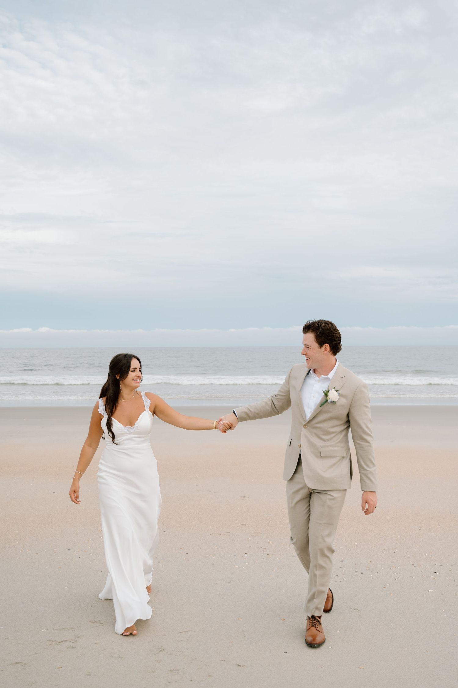 Bride and groom walk hand-in-hand on a sandy beach, she in a white gown, he in a tan suit.