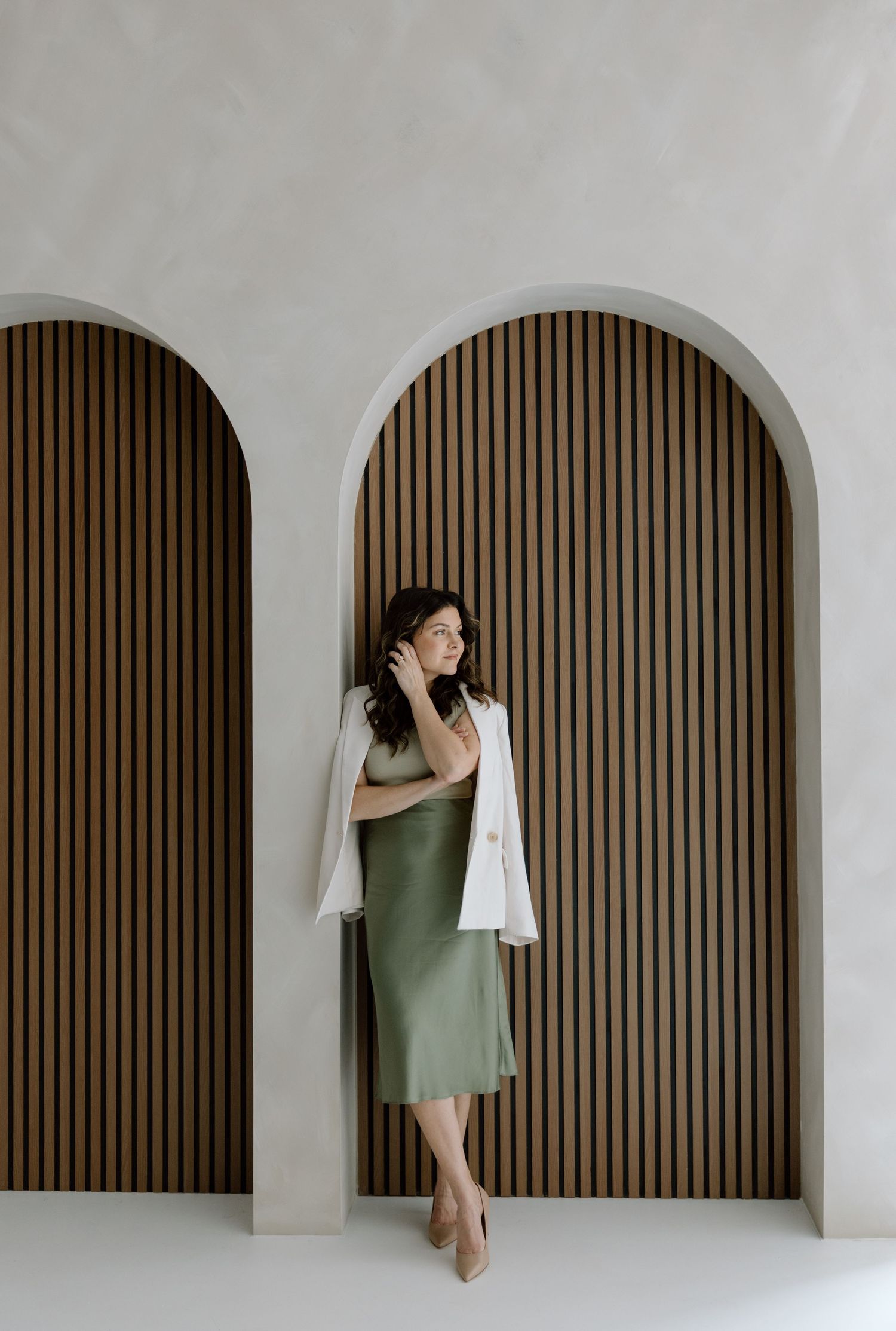A figure in an olive green dress and white shawl poses elegantly against a minimalist arched backdrop with wood slat details.