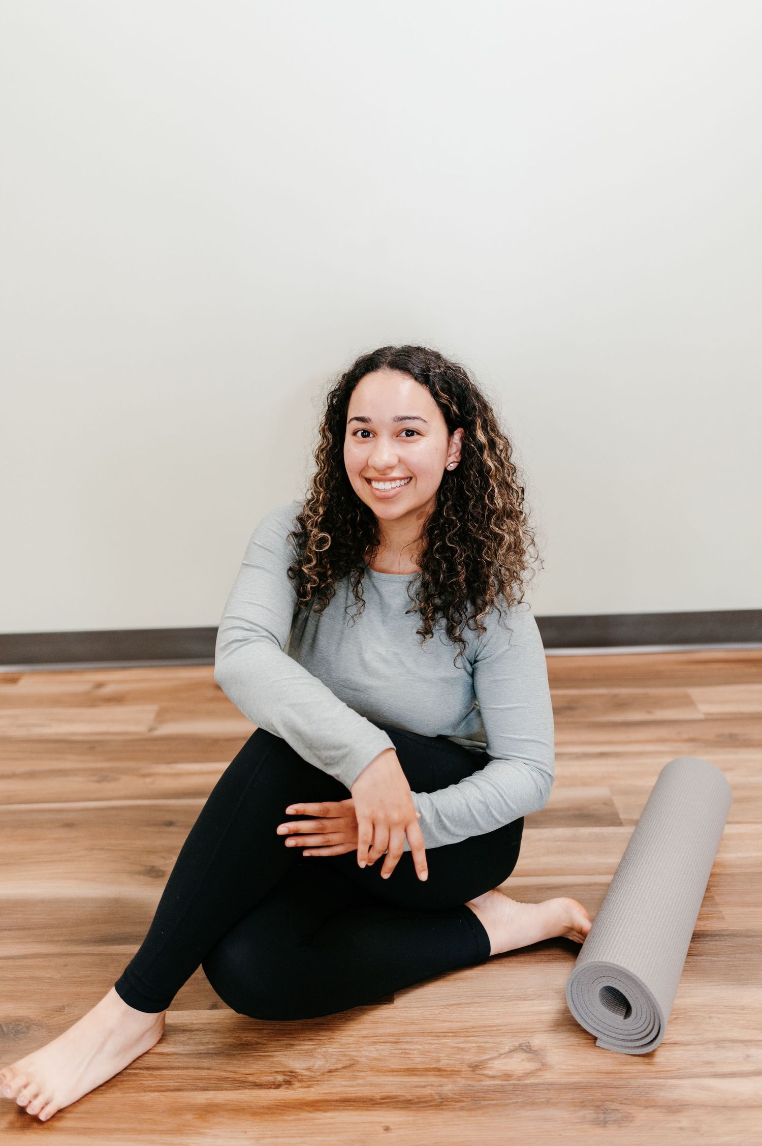 A person sits on wooden flooring in workout attire next to a rolled up gray yoga mat in a light-filled space.