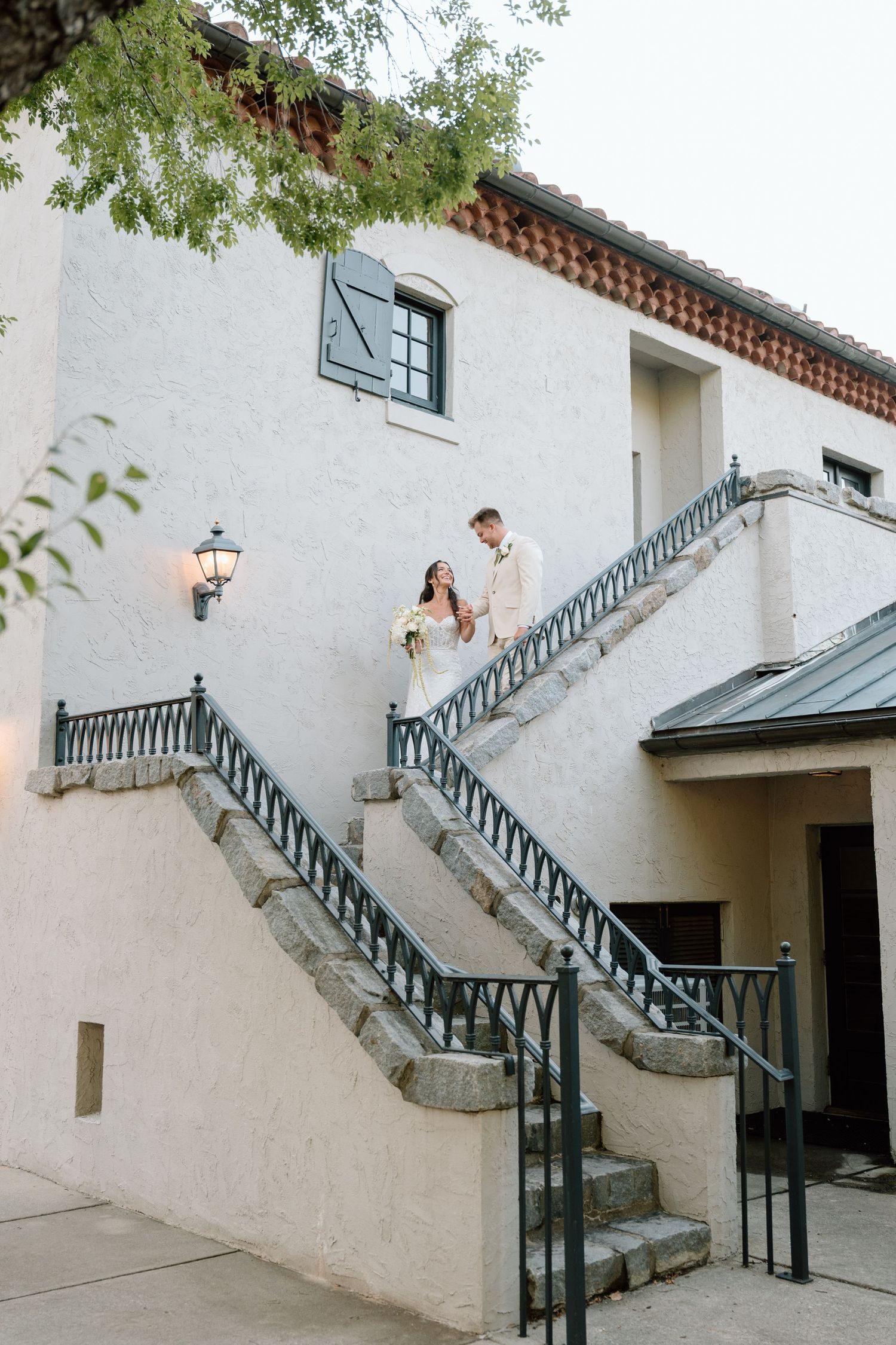 Bride and groom descend elegant stone staircase of white stucco wedding venue with wrought iron railings.