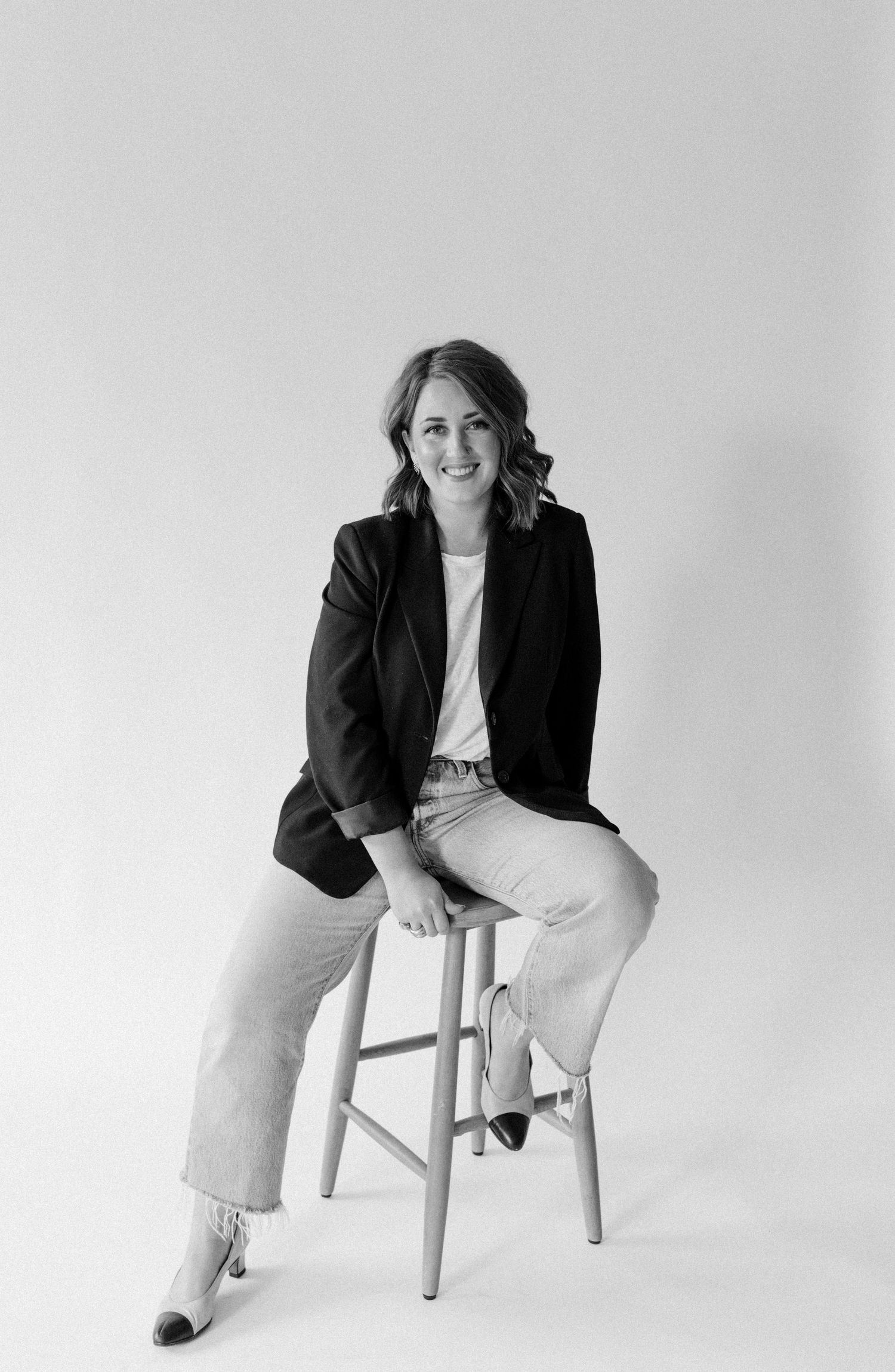 Professional black and white portrait of a person in a dark blazer and light pants sitting casually on a wooden stool.