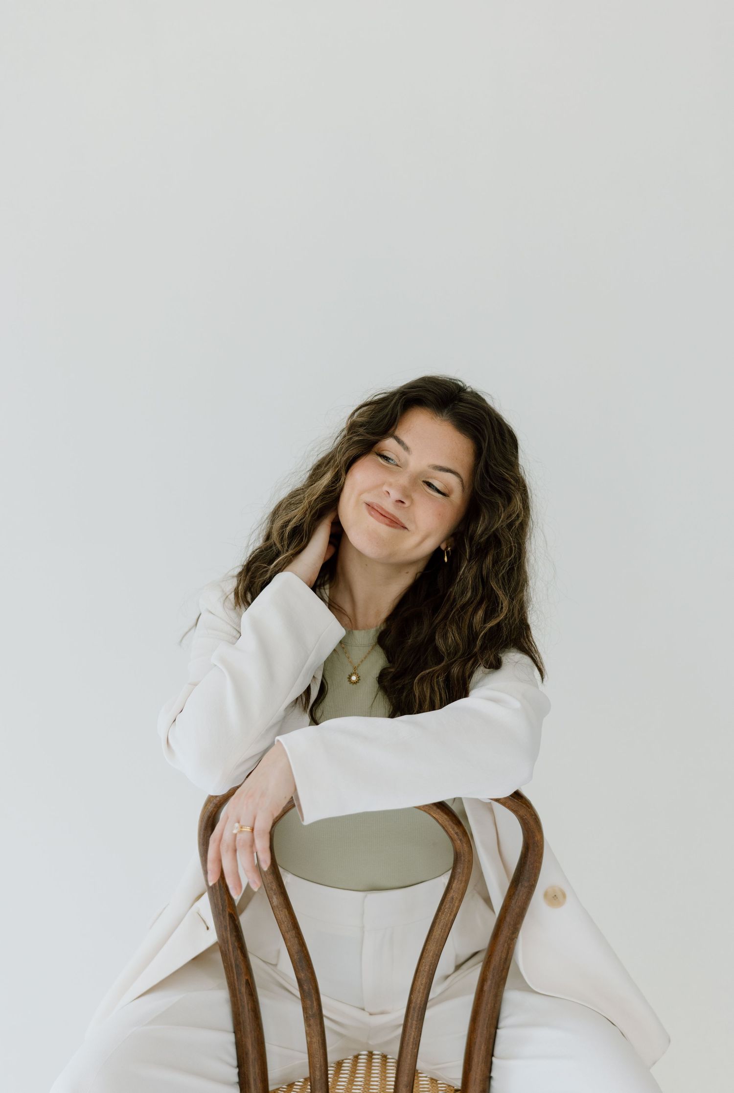 A person with long wavy hair sits casually on a white chair wearing a cream-colored outfit against a plain white wall.