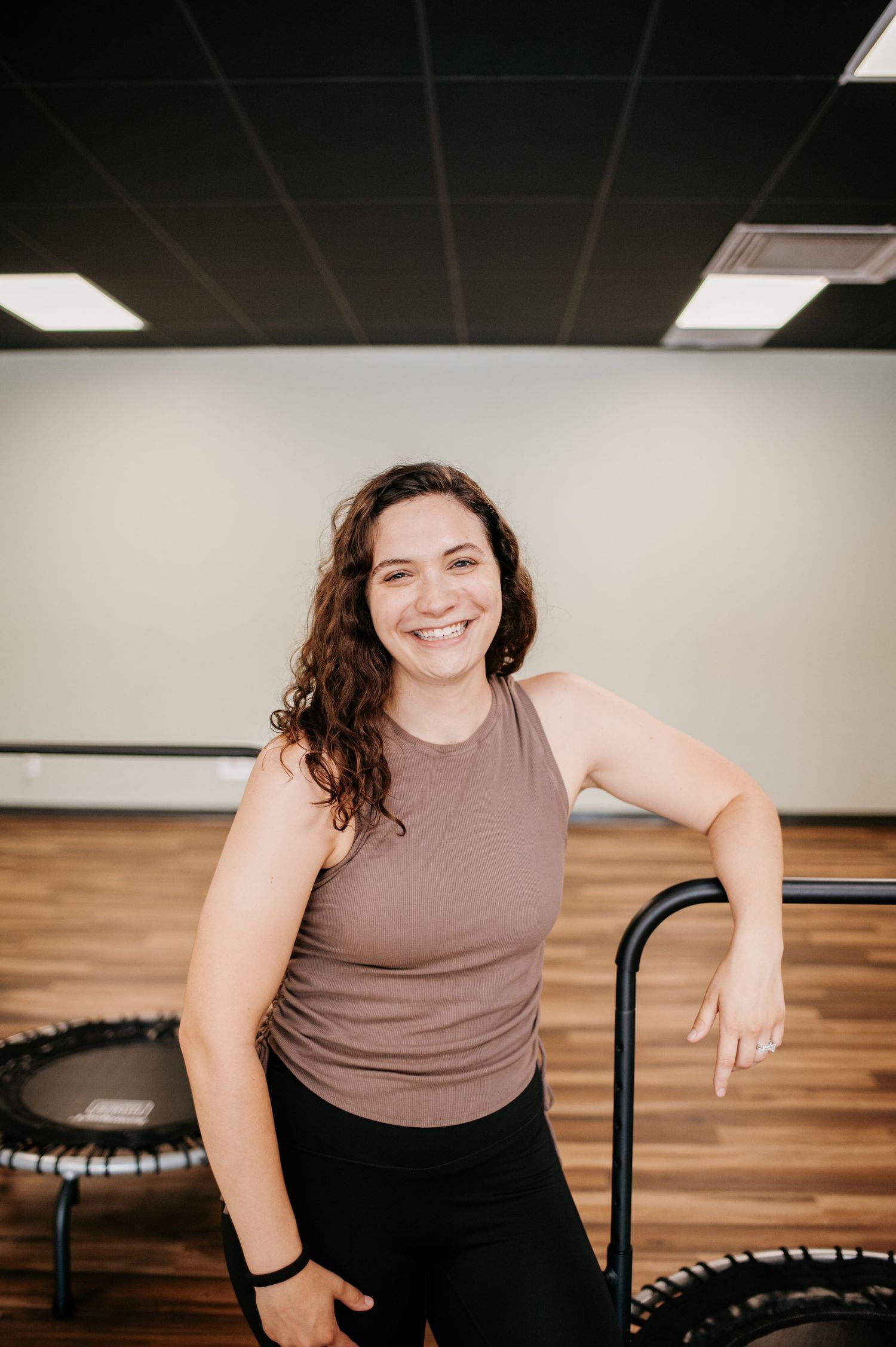 A fitness instructor in a mauve tank top smiles while standing next to a mini trampoline in an exercise studio.