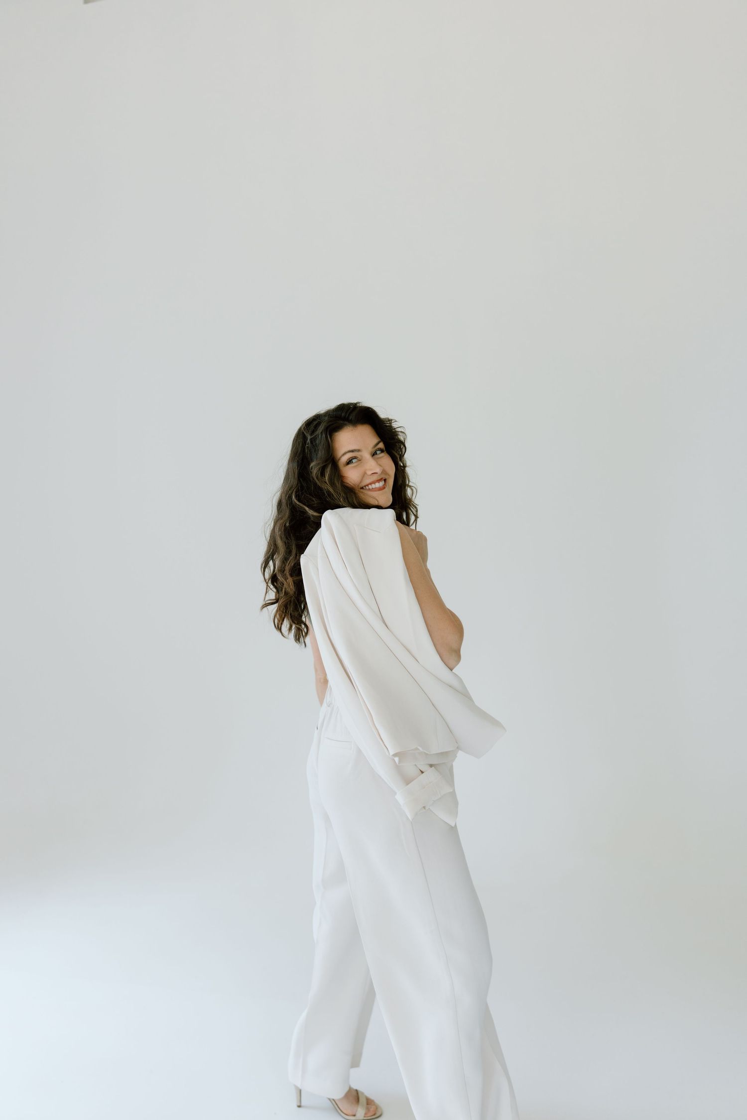 Figure in flowing white outfit turns playfully against minimal white background while long dark hair cascades.