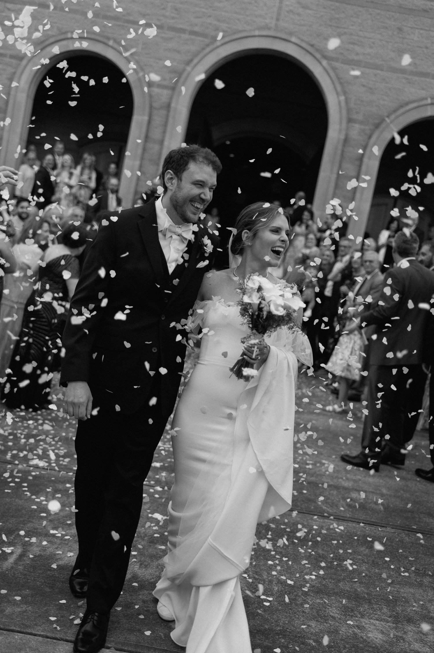 Black and white photo of a joyful bride and groom walking through confetti outside a church after their wedding ceremony.