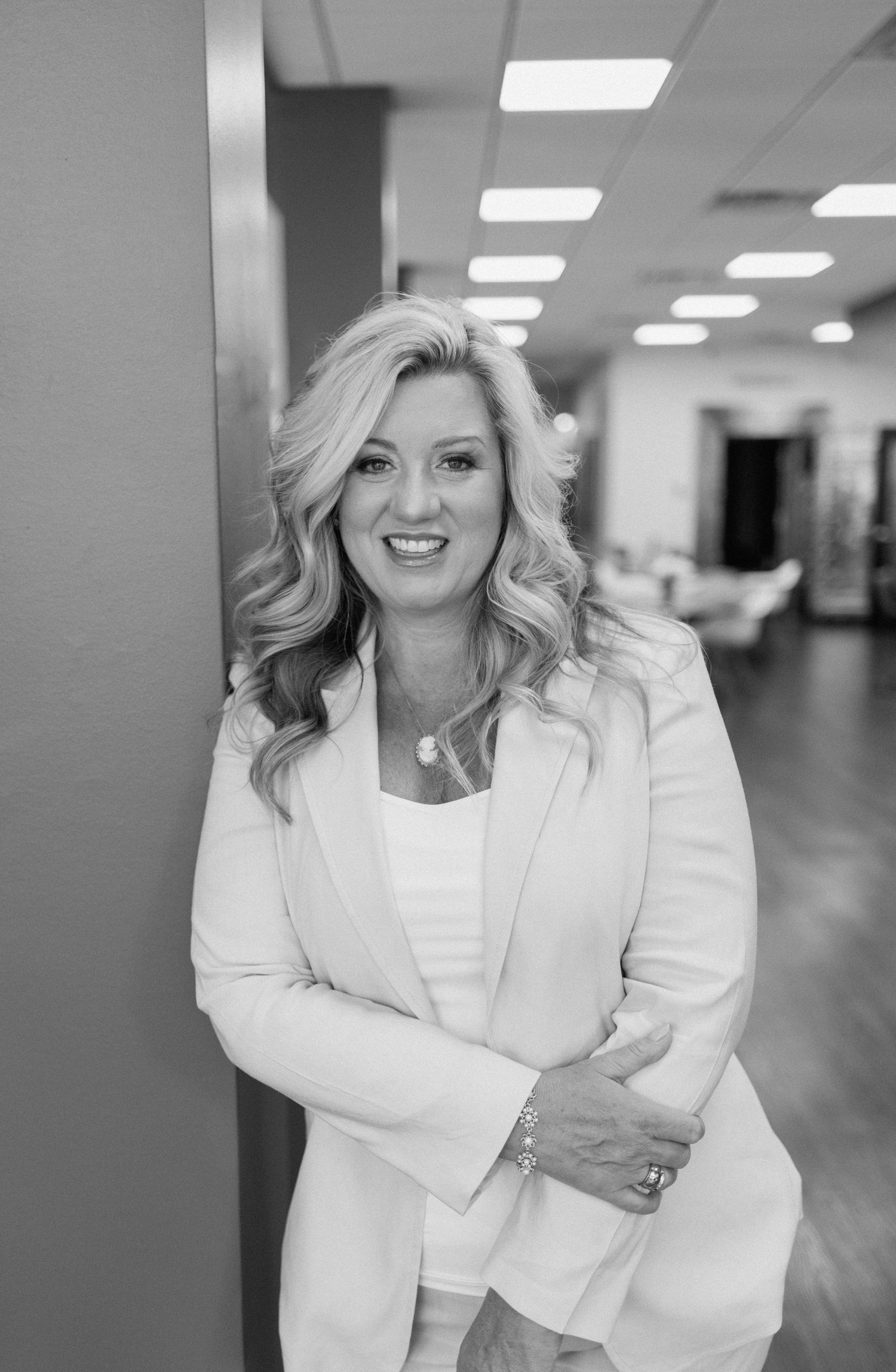 Professional black and white portrait of a person in a white blazer smiling in an office hallway.