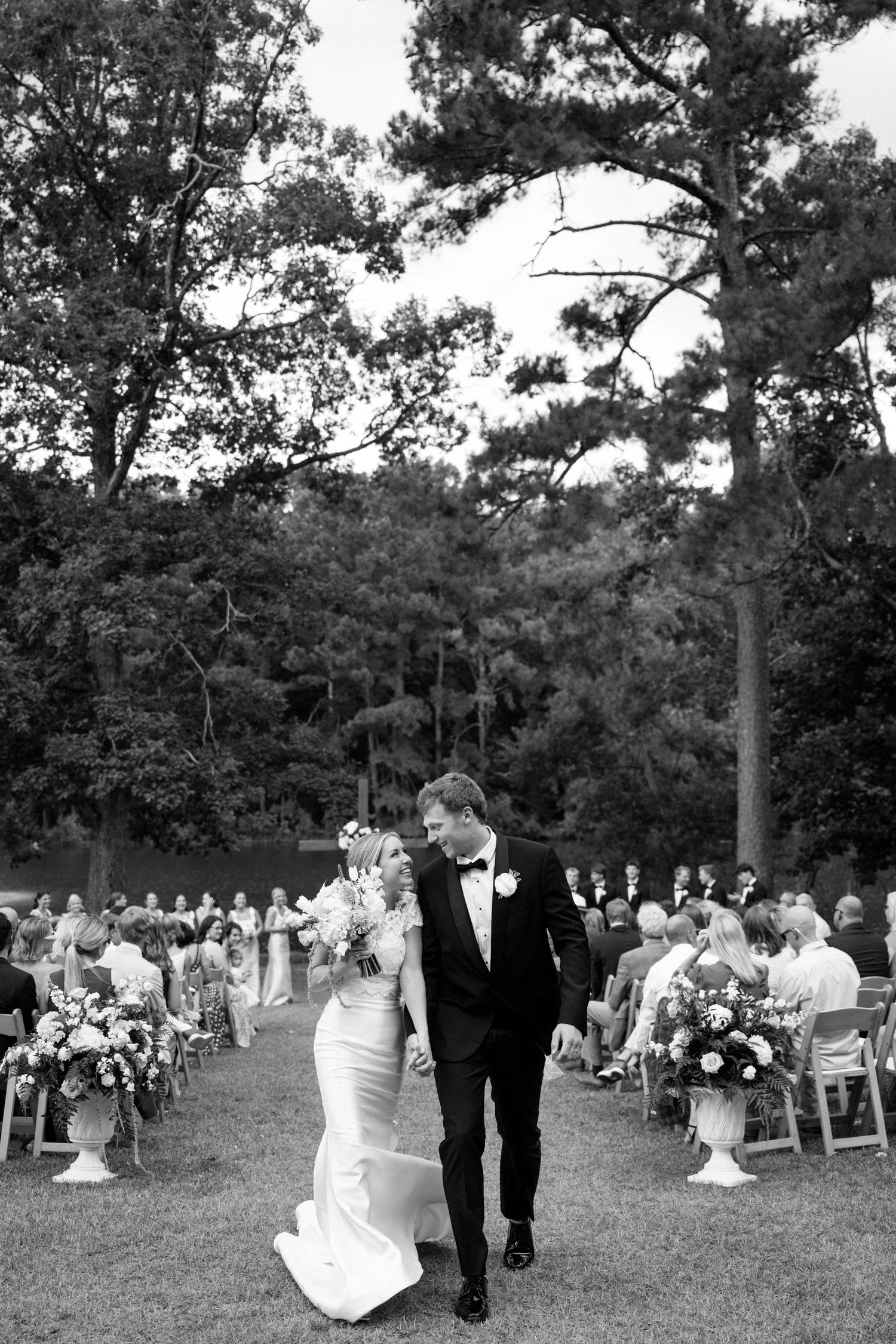 Black and white photo of bride and groom walking down outdoor aisle after wedding ceremony surrounded by guests.