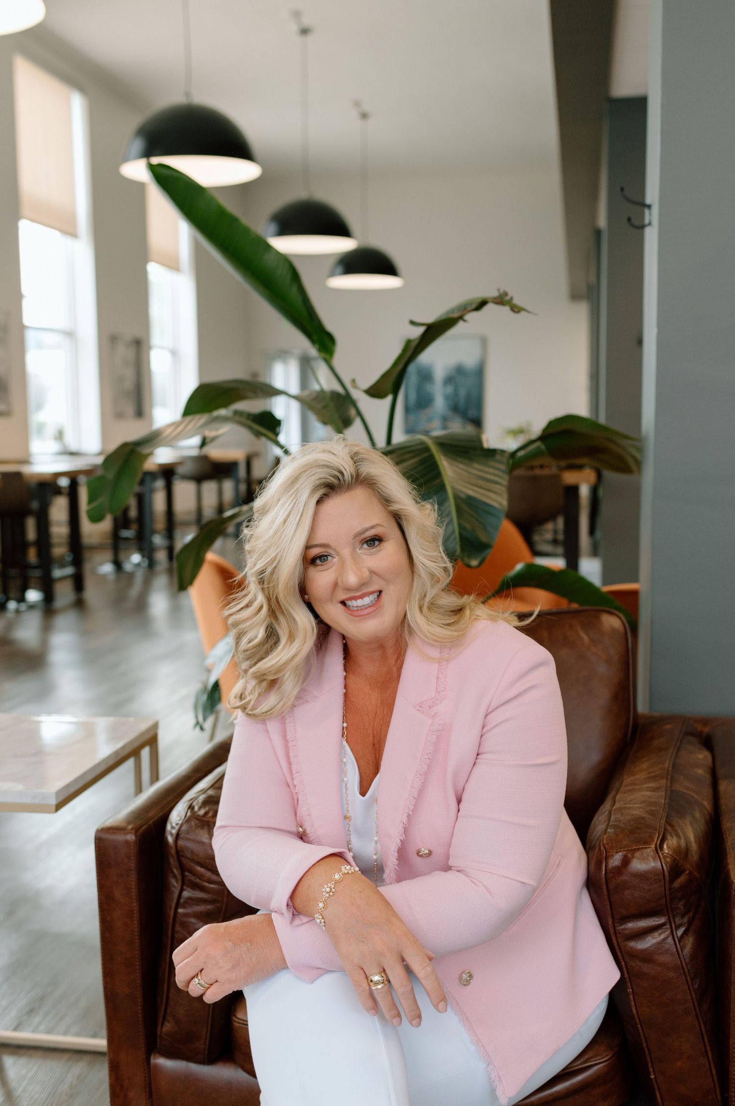 A person in a pink blazer sits on a brown leather chair in a modern office space with hanging pendant lights.