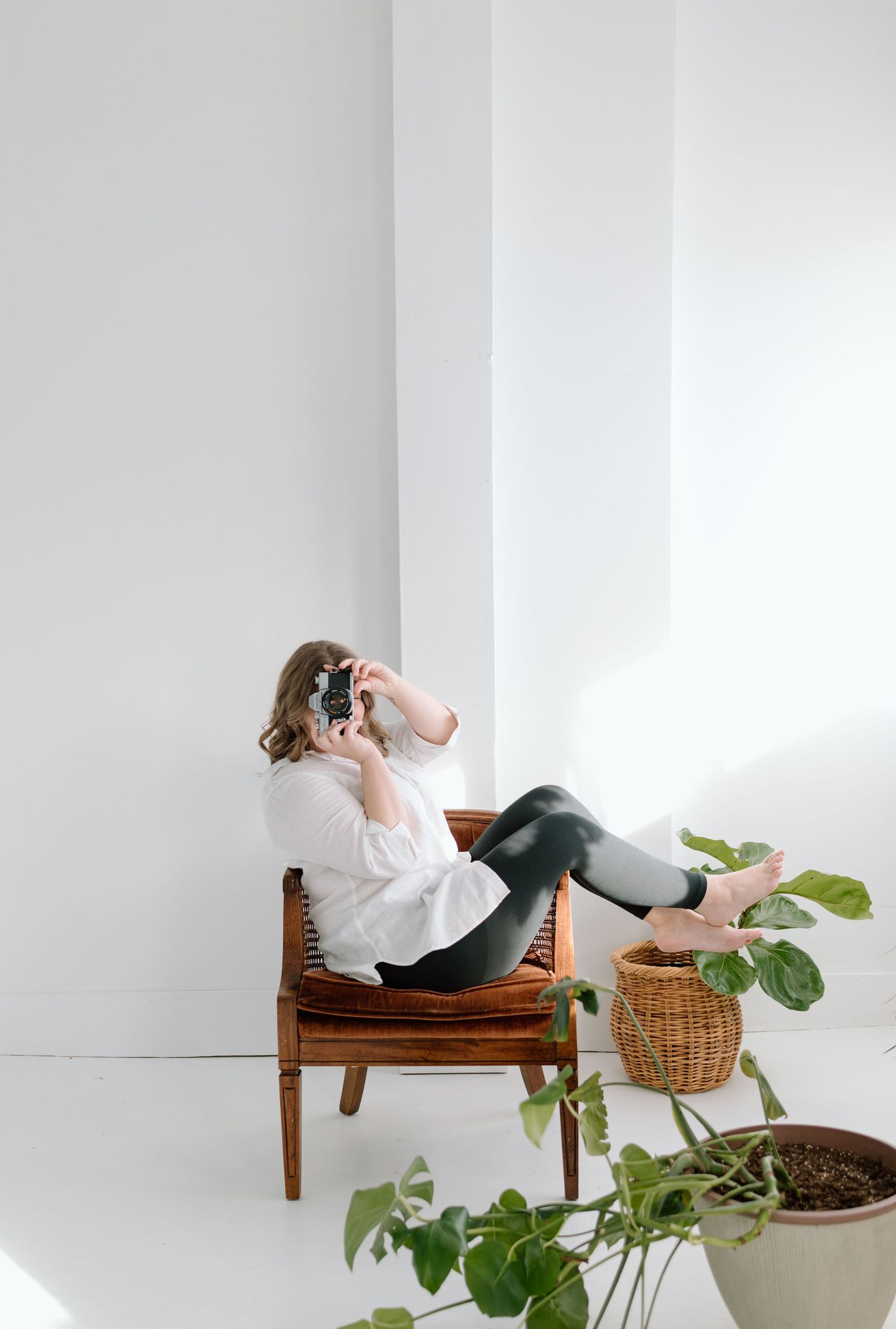 A person in a white top and leggings sits casually on a wooden chair with potted plants nearby in a bright minimal setting.