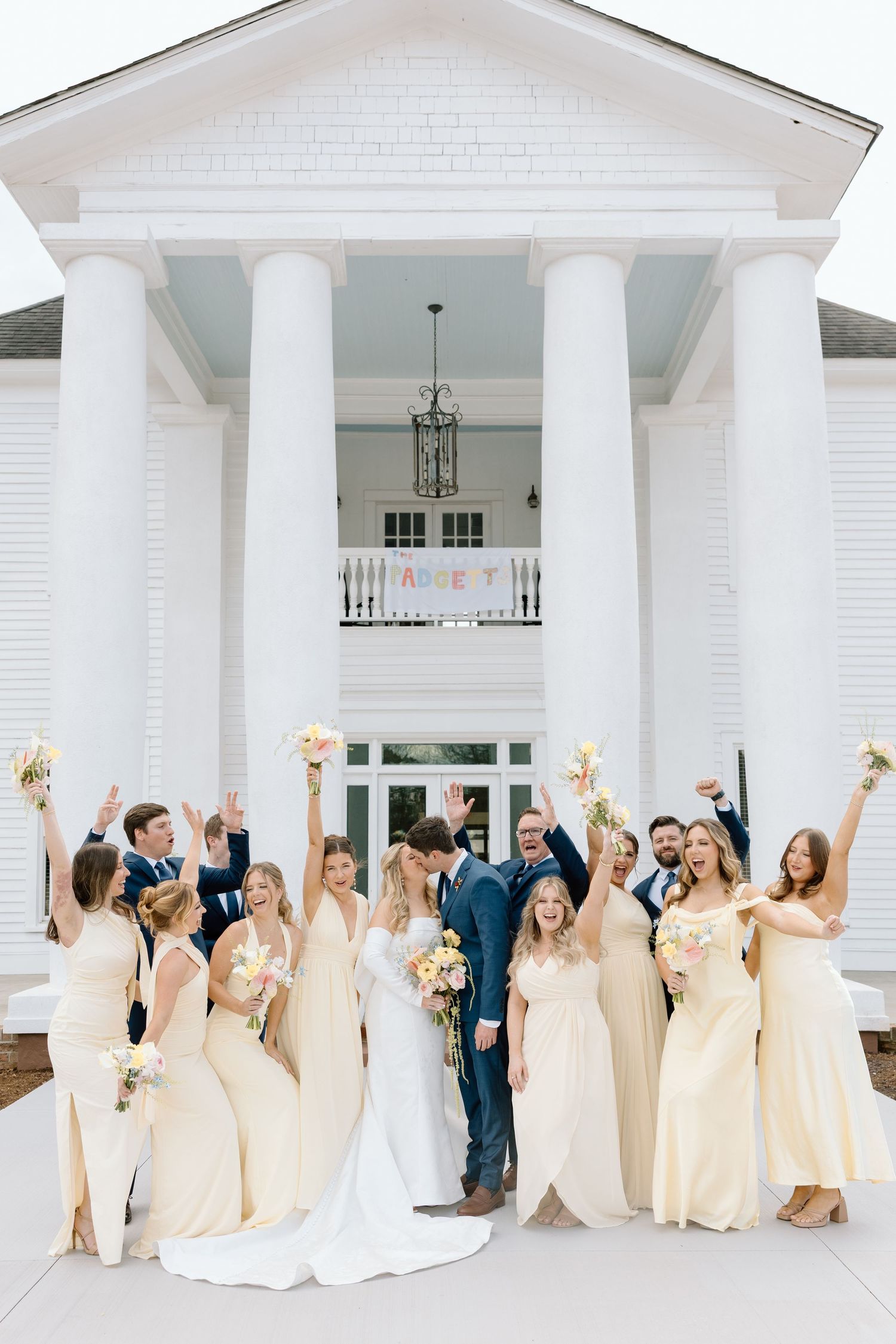 Wedding party celebrating in front of white colonial venue, bridesmaids in yellow gowns, arms raised in joy.