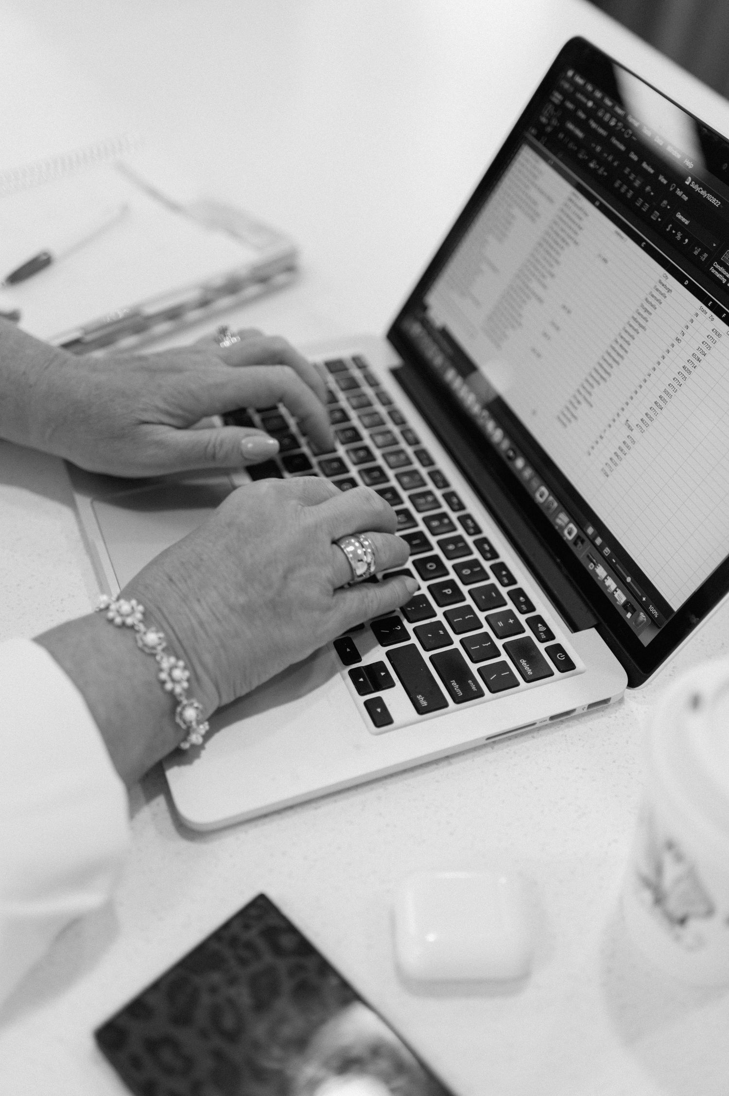 Black and white image of hands typing on a MacBook laptop keyboard with jewelry visible and spreadsheet on screen.