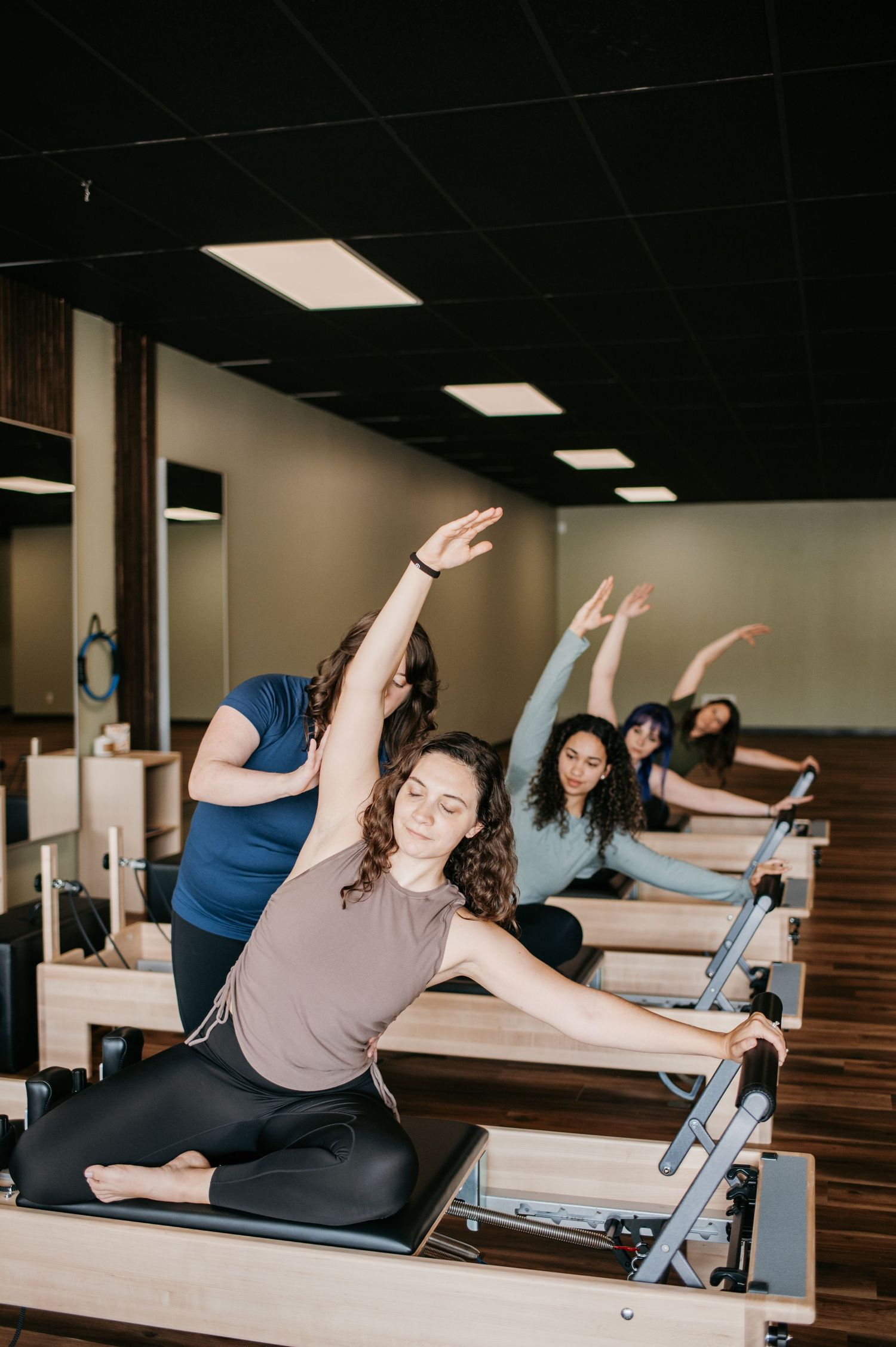 Students practice Pilates exercises on reformer machines in a modern fitness studio with overhead lighting.