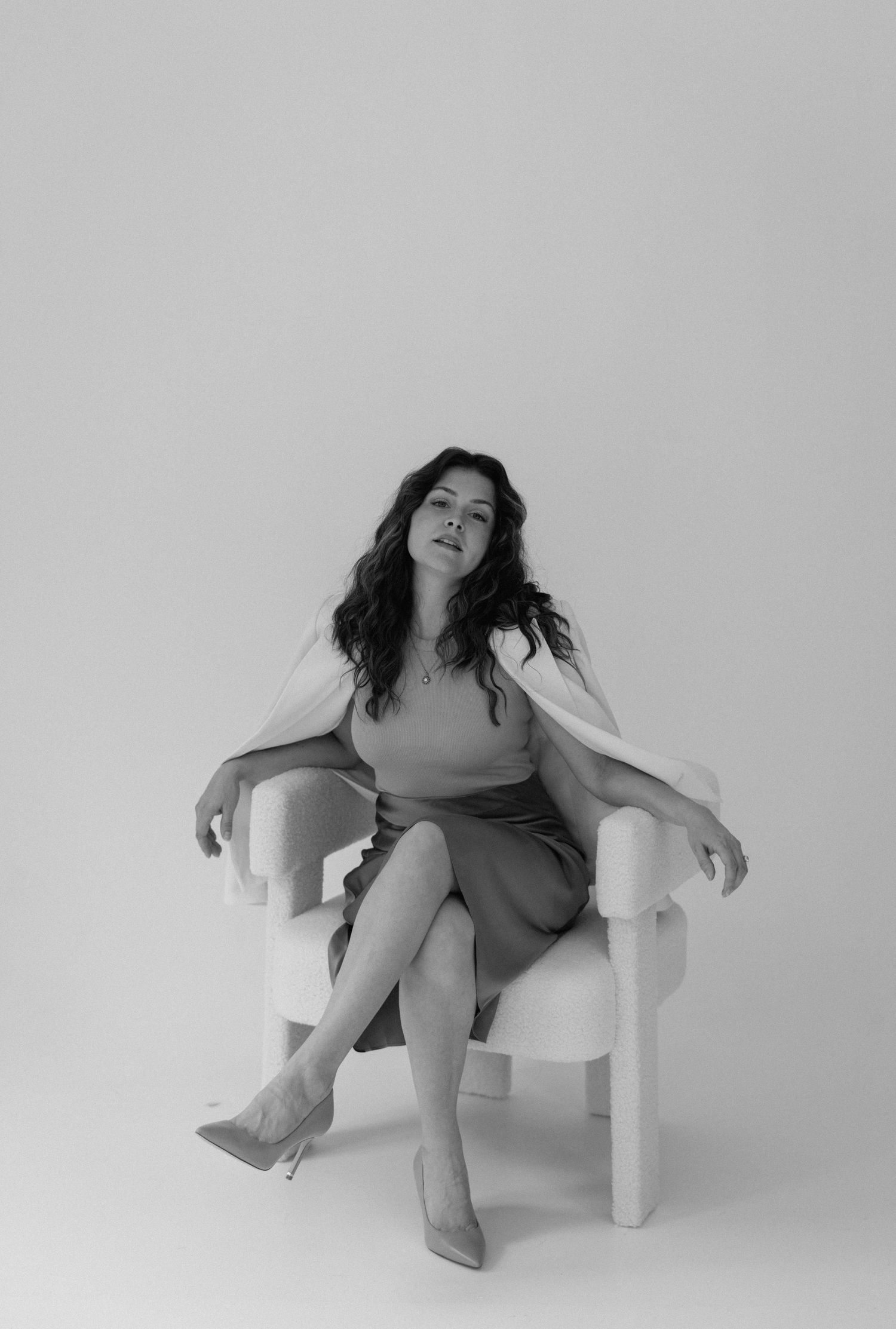 A black and white portrait of someone seated in a white chair wearing a fitted dress and heels in a minimalist setting.
