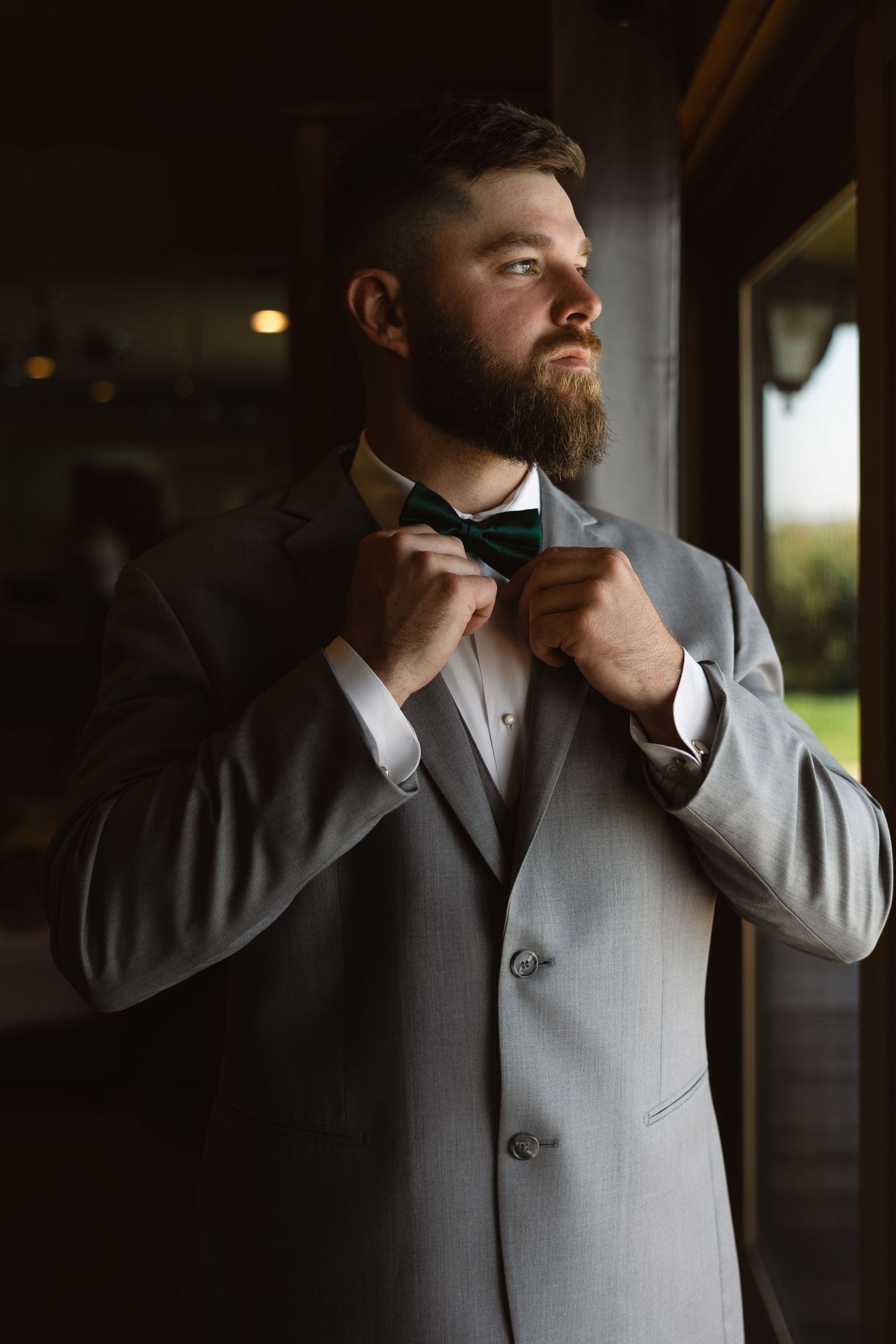 A person in formal attire adjusts their black bow tie while wearing a light gray suit jacket in moody lighting.
