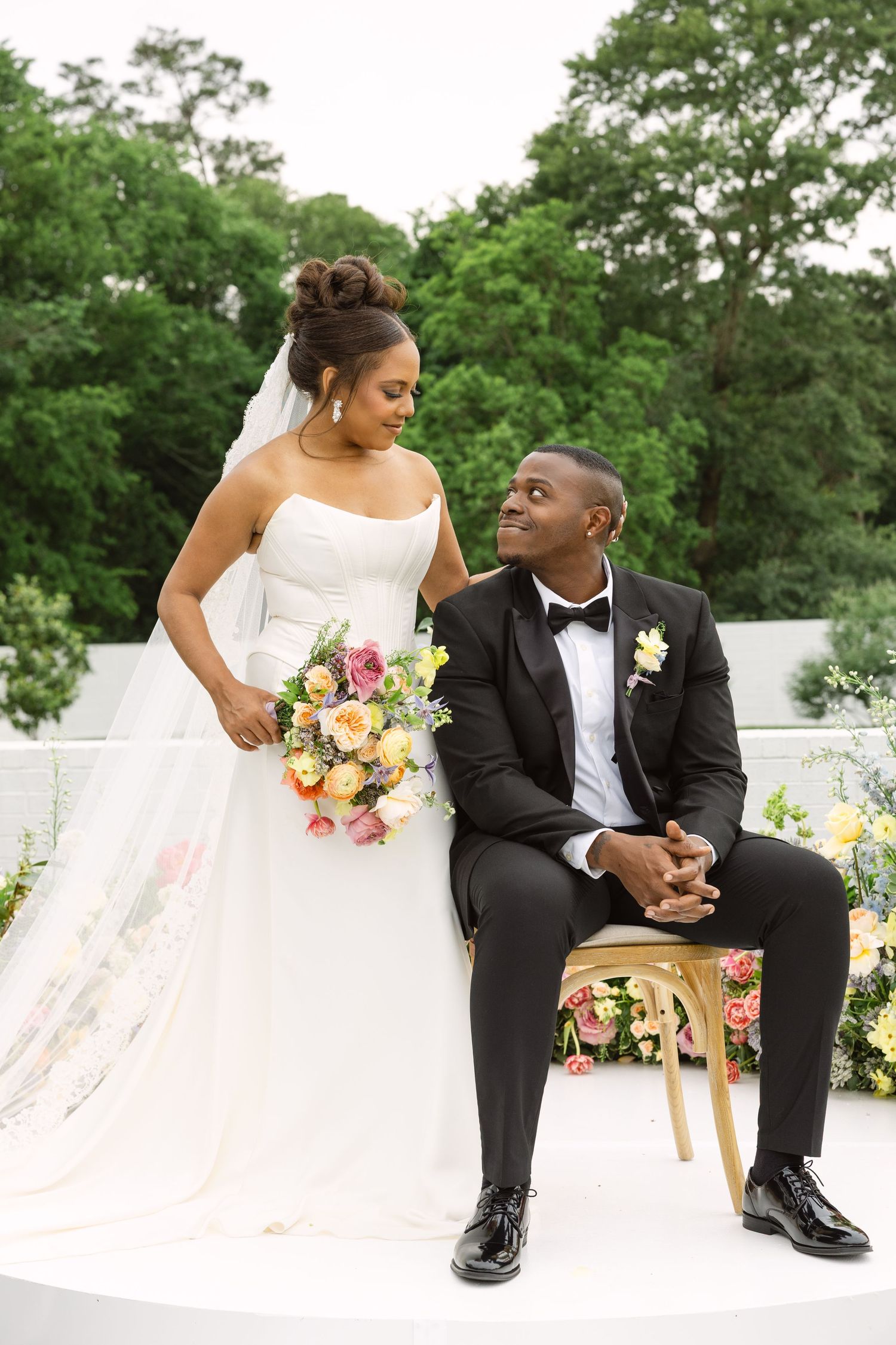 Elegant couple at outdoor wedding ceremony with bride holding colorful bouquet and groom seated on gold chair.