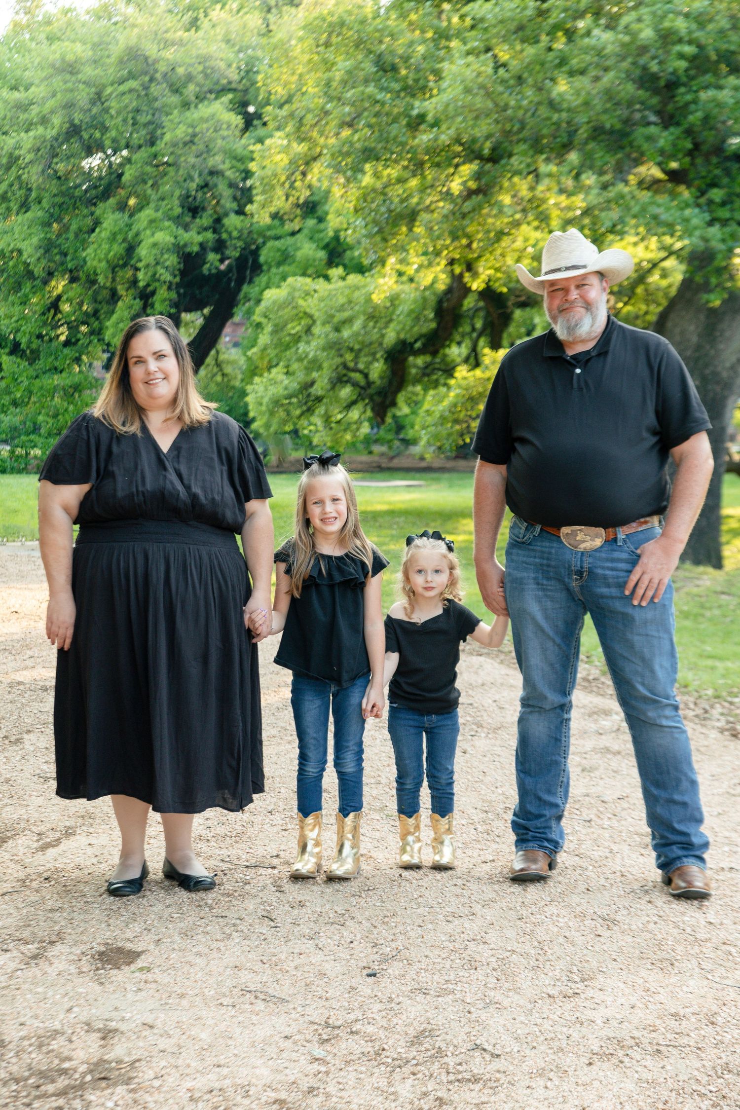 A family poses together on a dirt path surrounded by lush green trees in a park setting on a sunny day.